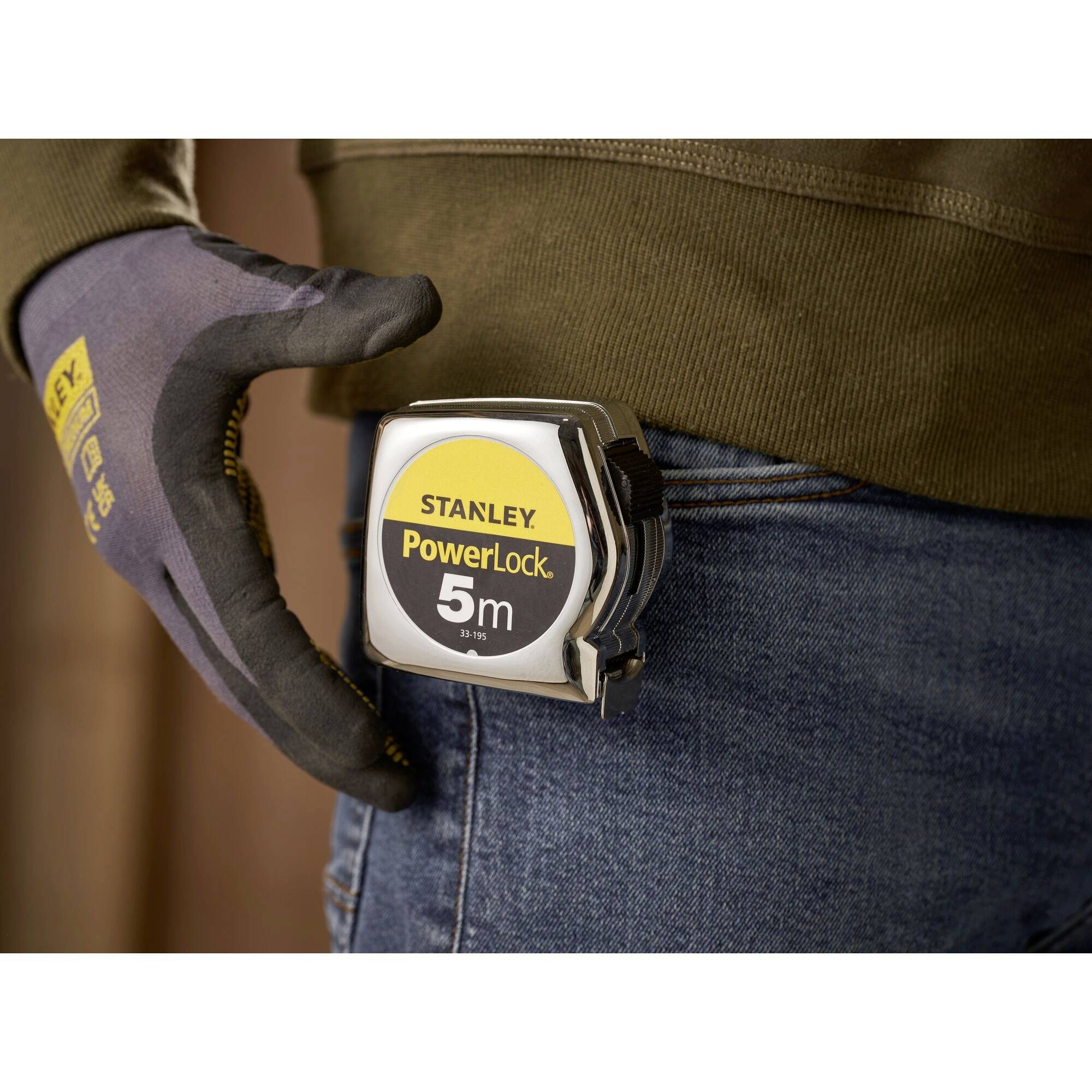Hand hält ein 5-Meter-Stanley-Maßband an Gürtel. Gezeigt wird funktionale Nutzung bei der Arbeit mit Handschuhen.