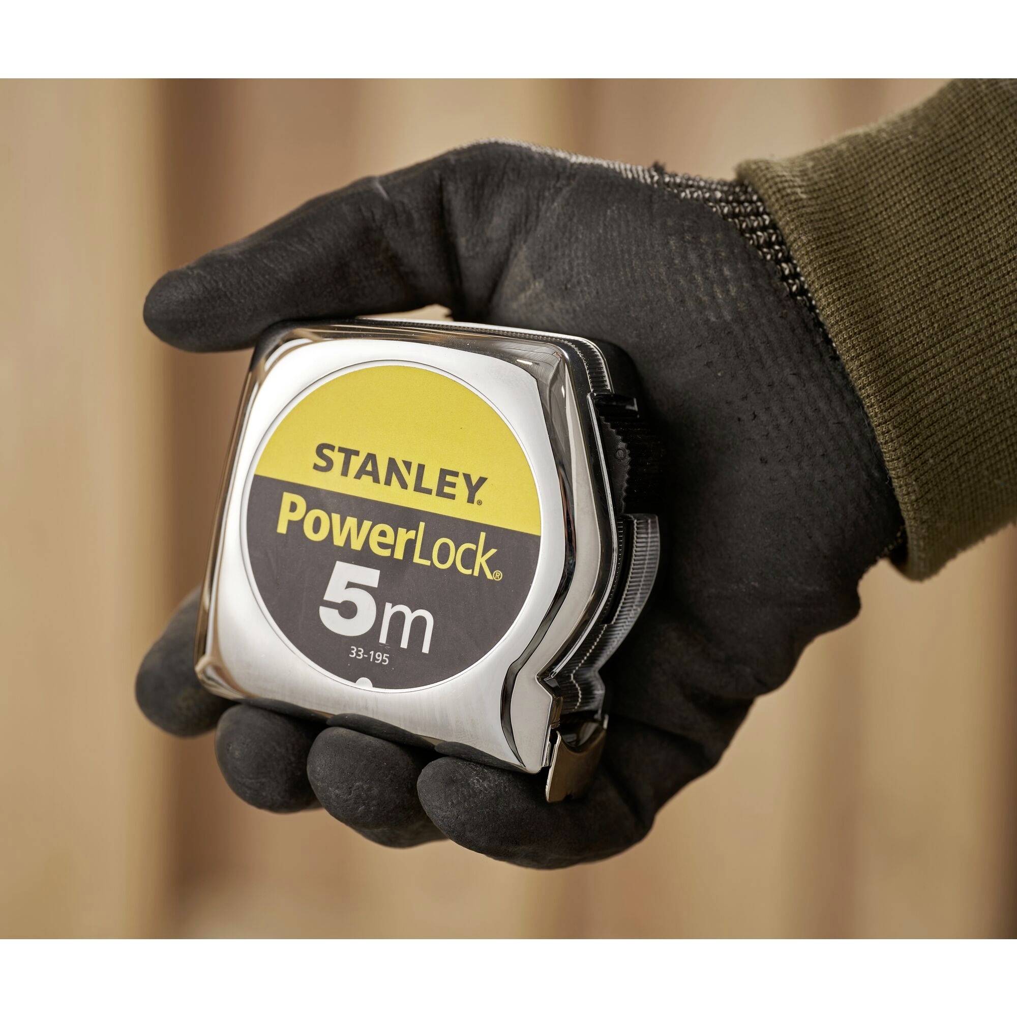 Ein Handschuh hält ein Maßband der Marke Stanley mit der Aufschrift 'PowerLock 5m', vor einem unscharfen Hintergrund.