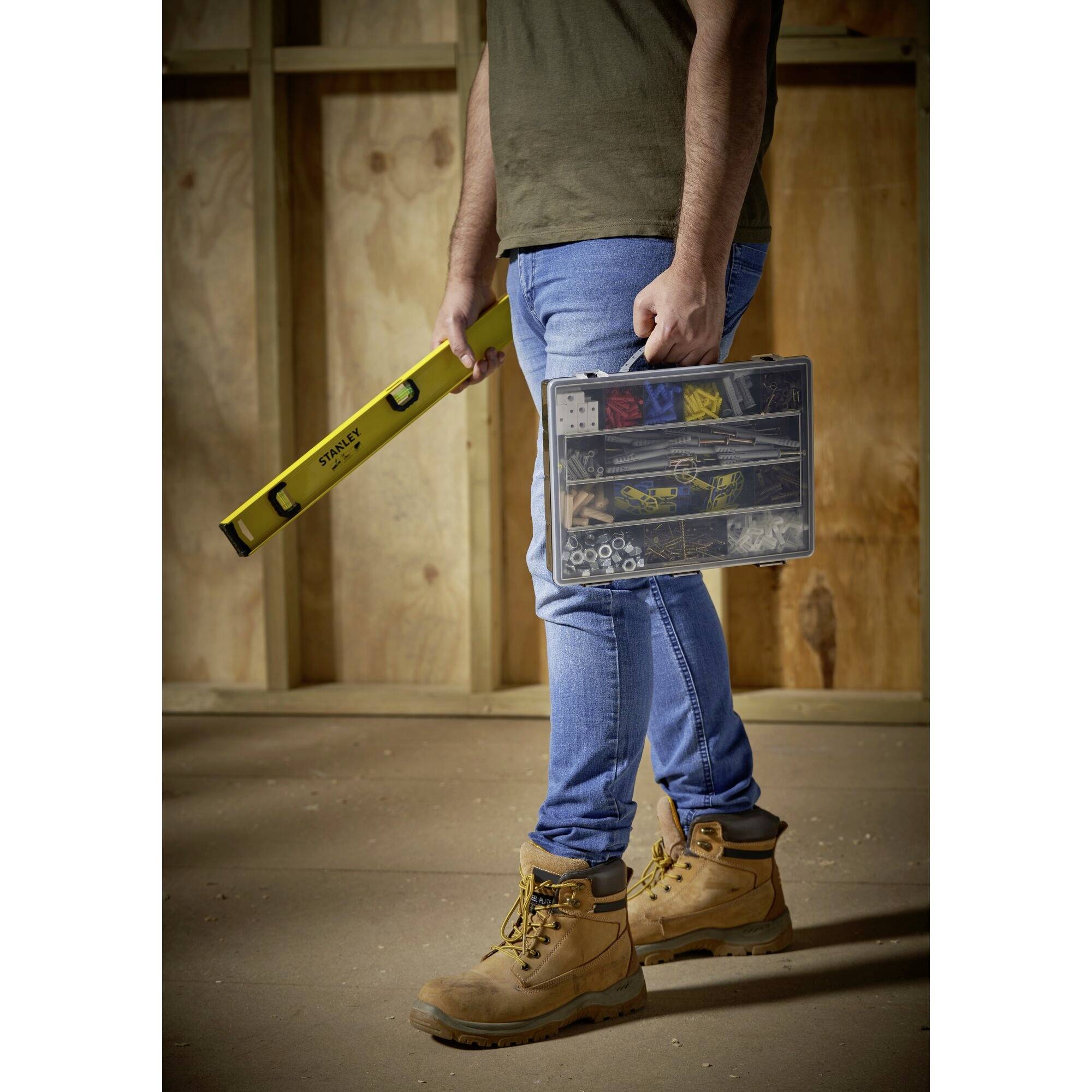 Ein Mann in Jeans und braunen Stiefeln hält eine Wasserwaage und eine Kiste mit Schrauben und Nägeln in einem Holzraum.