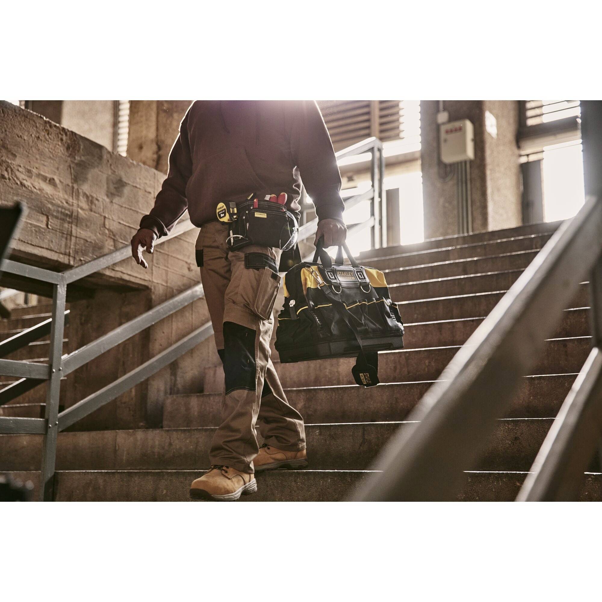 Eine Person in Arbeitskleidung, trägt eine Werkzeugtasche und geht eine Treppe hinunter. Umgebung erscheint industriell.