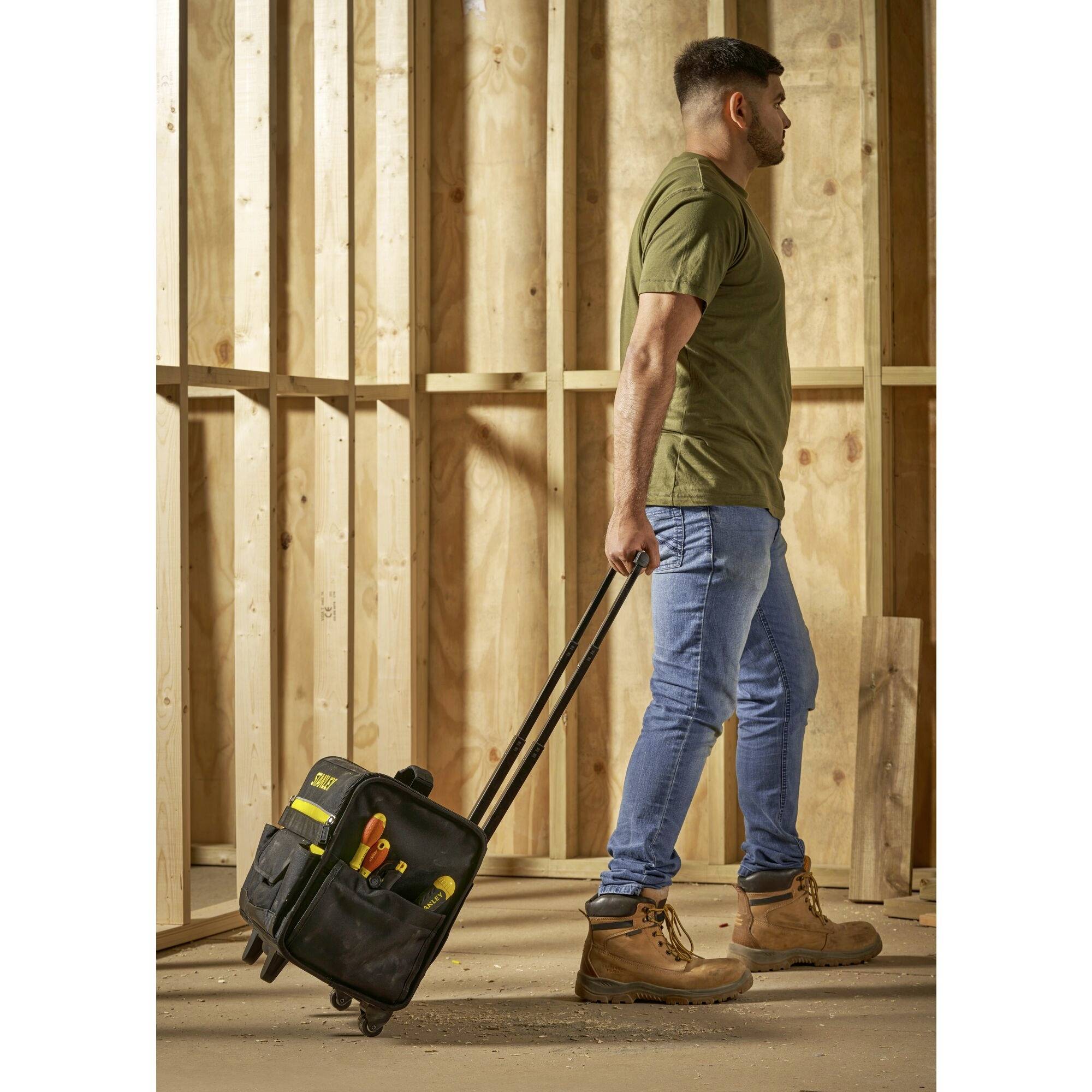 Eine Person zieht eine schwarze Werkzeugtasche mit Rädern auf einer Baustelle. Im Hintergrund sind Holzwände sichtbar.