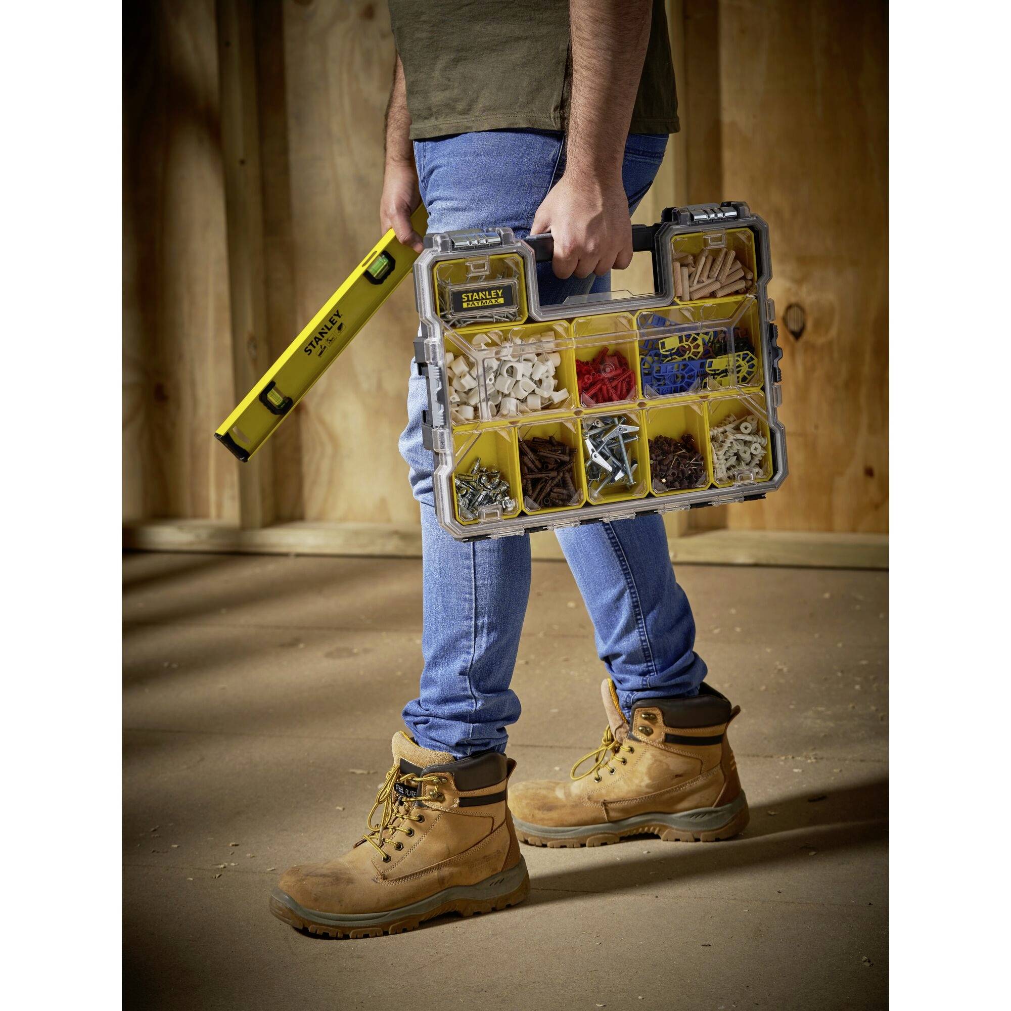 Eine Person trägt eine gelbe Wasserwaage und einen klaren Werkzeugkoffer mit verschiedenen Schrauben und Dübeln, während sie in einem unfertigen Raum steht.