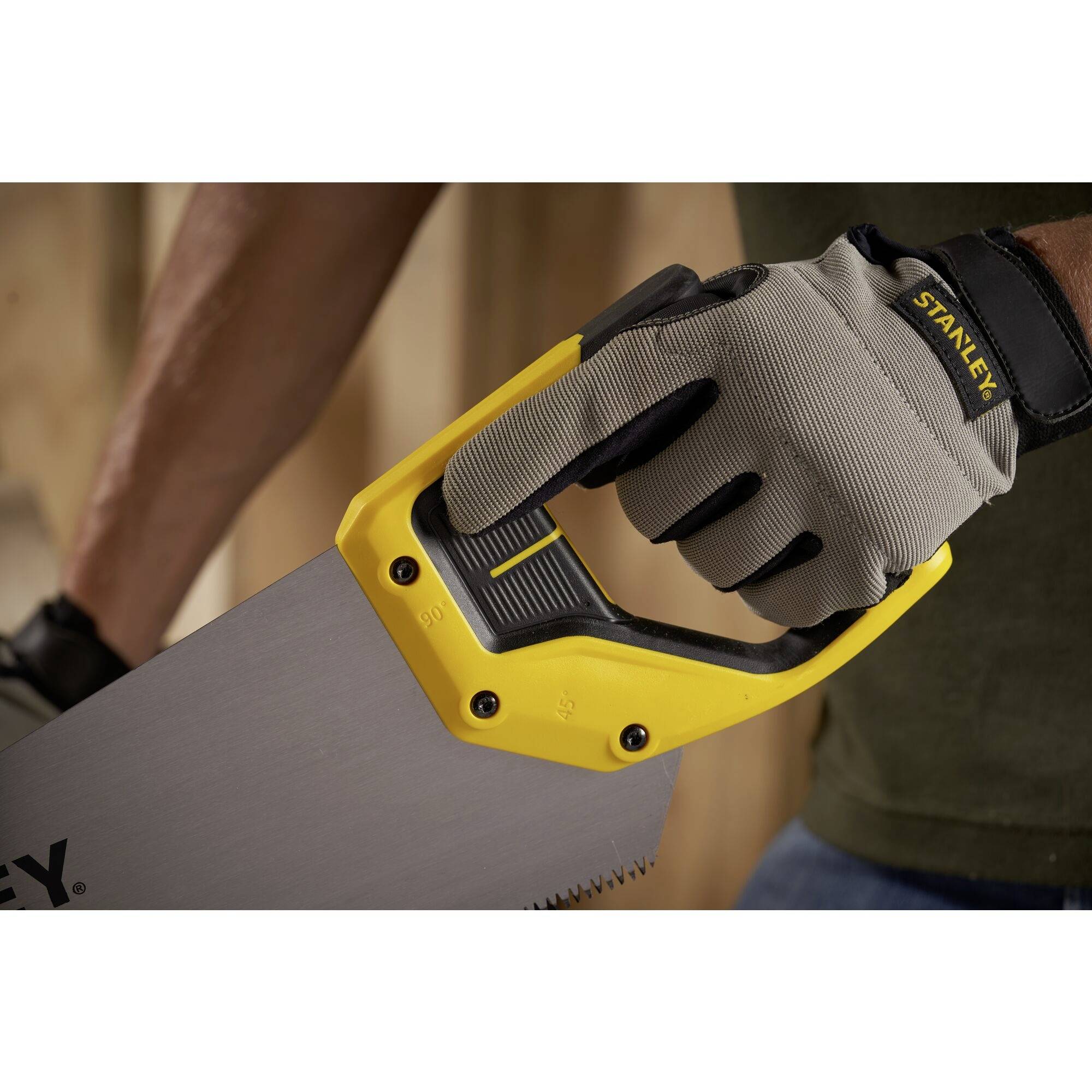 Eine Person trägt einen Handschuh und hält eine Handsäge mit gelbem Griff. Sie steht vor einer Holzwand, bereit zum Schneiden.