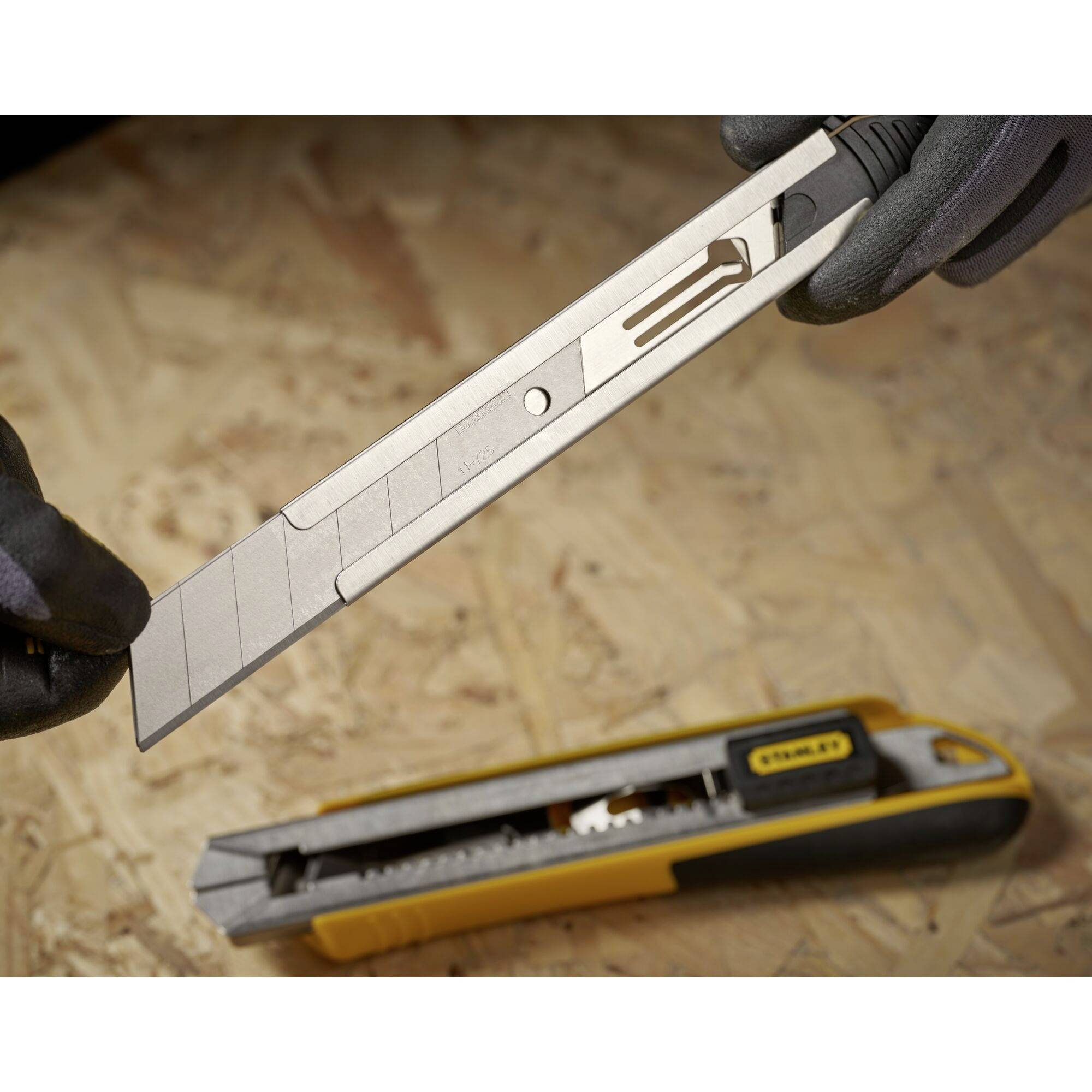 Eine Person mit Handschuhen wechselt die Klinge eines gelben Cuttermessers auf einer Holzwerkbank.
