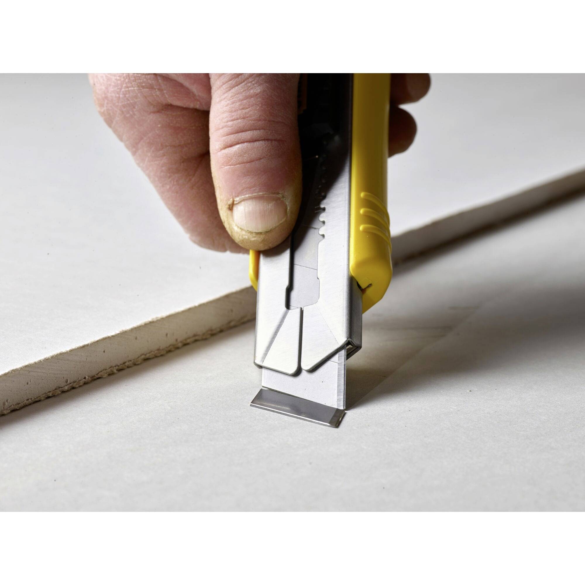 Eine Hand schneidet mit einem gelben Cutter-Messer ein Stück Gipskarton. Der Schnitt verläuft gerade entlang einer Linie.