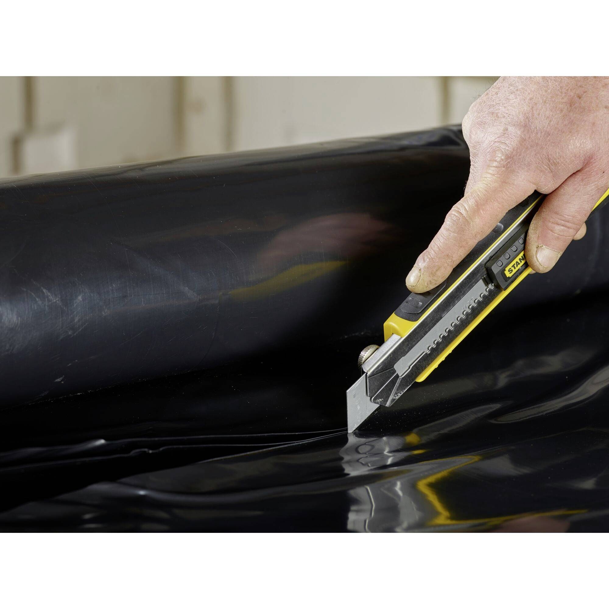 Eine Hand schneidet mit einem gelben Cuttermesser durch eine schwarze Plastikfolie.