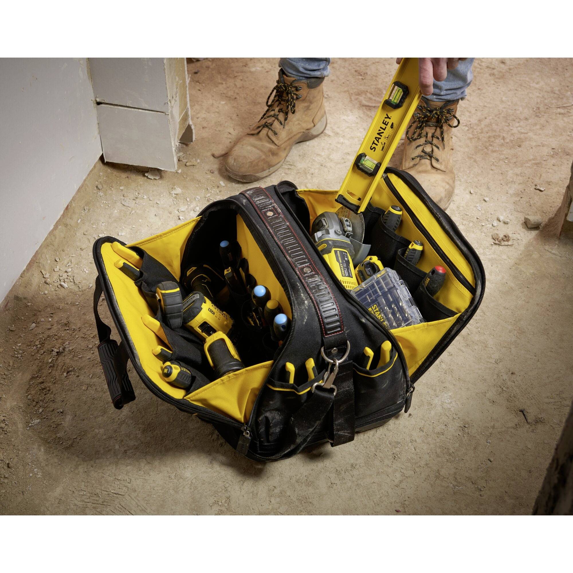 Eine geöffnete Werkzeugtasche auf einem Boden, gefüllt mit verschiedenen Werkzeugen wie Bohrmaschine und Schraubendrehern. Eine Person in Arbeitskleidung greift hinein.