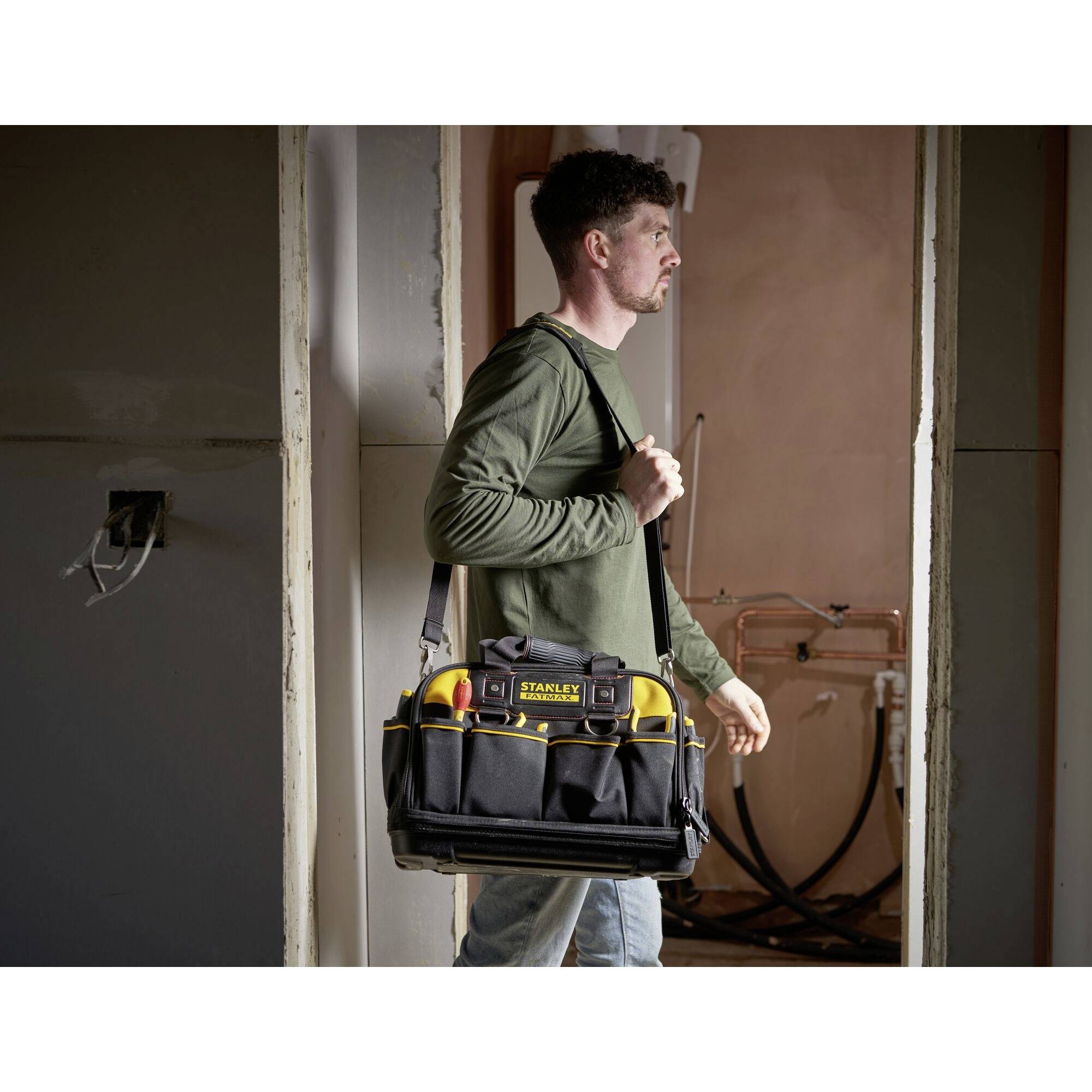 Ein Mann in einem Raum trägt eine große Werkzeugtasche auf der Schulter, bereit für handwerkliche Arbeiten. Im Hintergrund ist ein Badezimmer sichtbar.