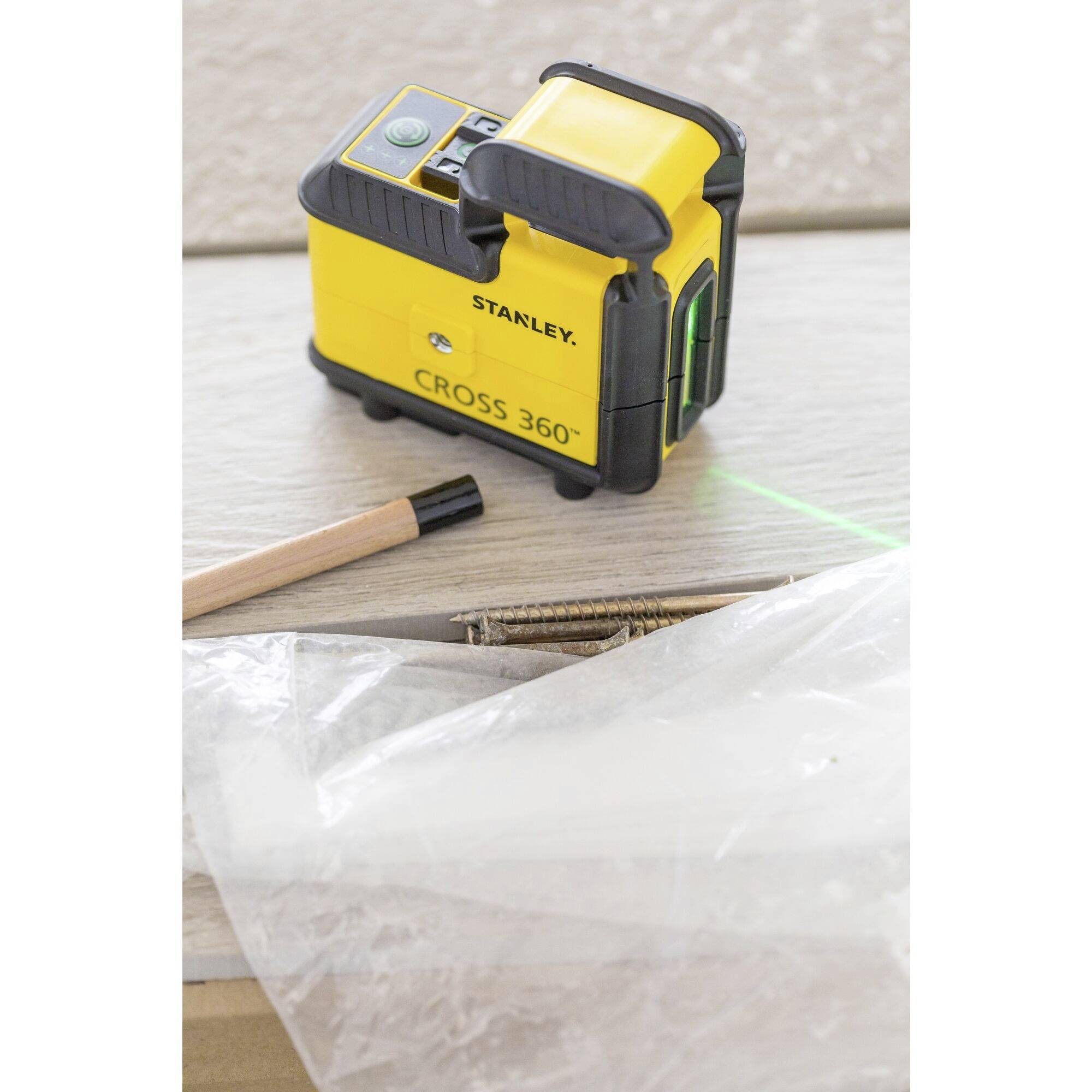 Ein gelbes Stanley Cross 360° Laser-Level-Werkzeug auf einem Holztisch, umgeben von Plastikfolie und einem Bleistift.