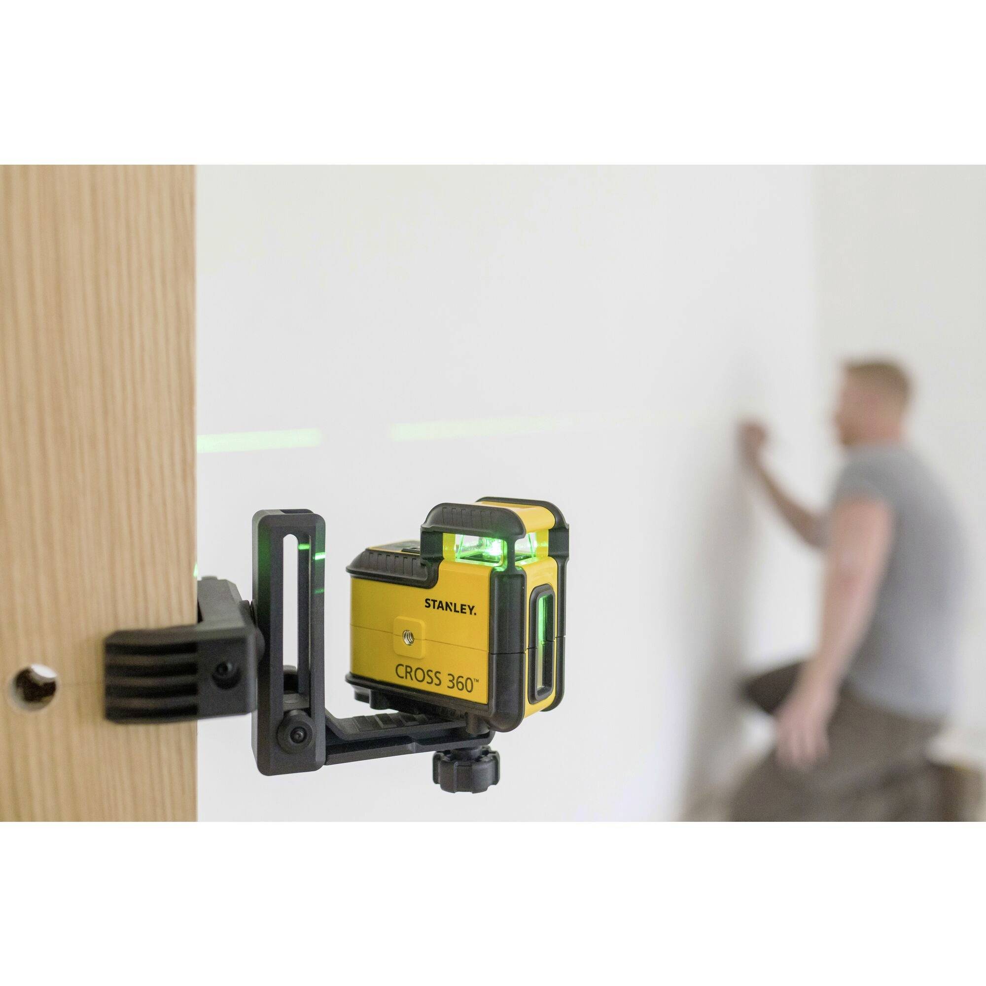 Ein Kreuzlinienlaser ist an einem Holzstück befestigt und projiziert grüne Linien auf eine weiße Wand. Im Hintergrund arbeitet eine unscharfe Person an der Wand.