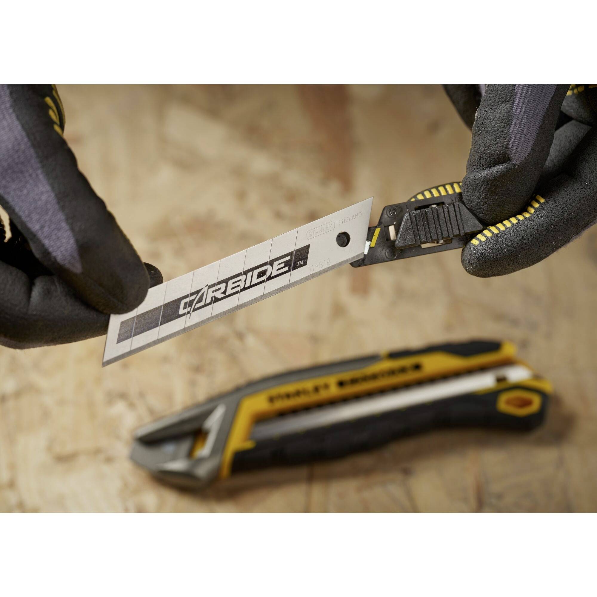 Zwei behandschuhte Hände halten eine Klinge mit der Aufschrift 'Carbide', im Hintergrund ein gelbes Cuttermesser auf Holzuntergrund.