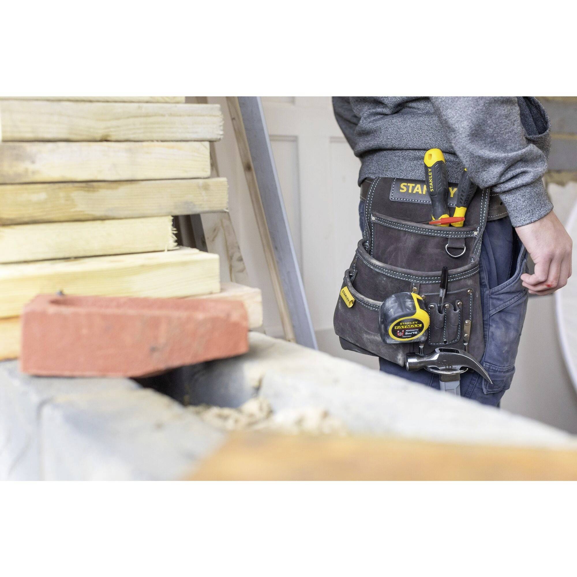 Ein Handwerker mit Werkzeugtasche steht neben einem Stapel Holz. Ein Zollstock und ein Schraubendreher sind sichtbar in der Tasche.