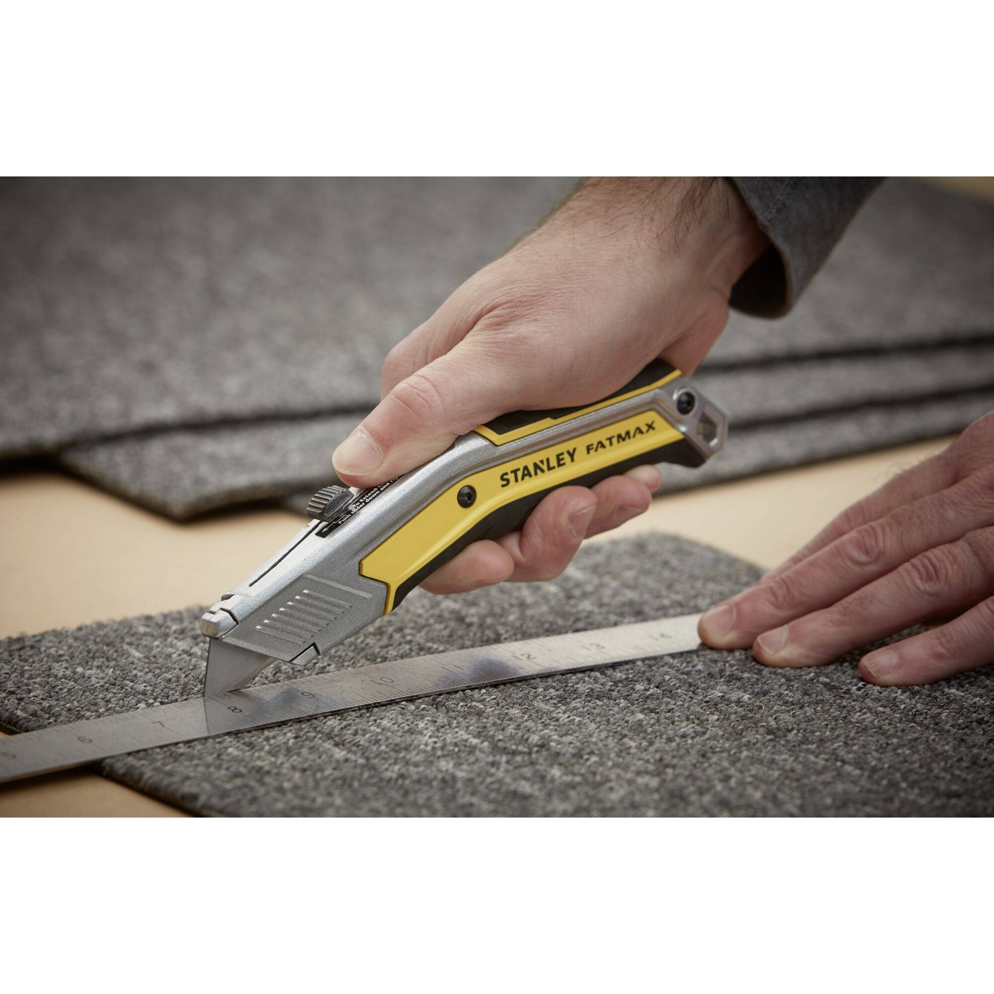 Eine Hand schneidet mit einem gelben Stanley-FatMax-Messer, unterstützt von einem Metalllineal, präzise grauen Teppichboden.