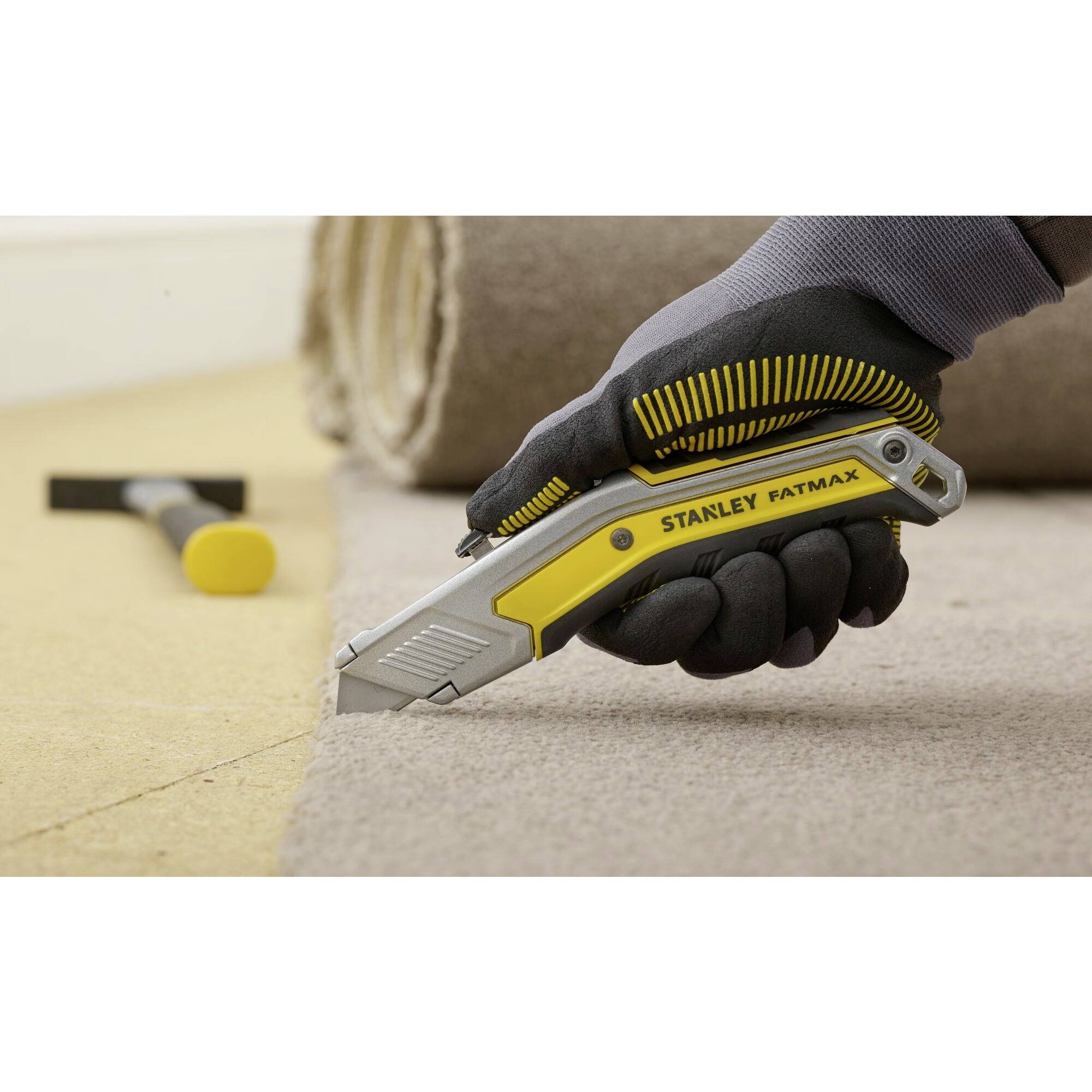 Eine Hand mit Handschuh schneidet mit einem gelben Stanley FatMax Cuttermesser durch Teppichboden. Im Hintergrund ein Hammer.