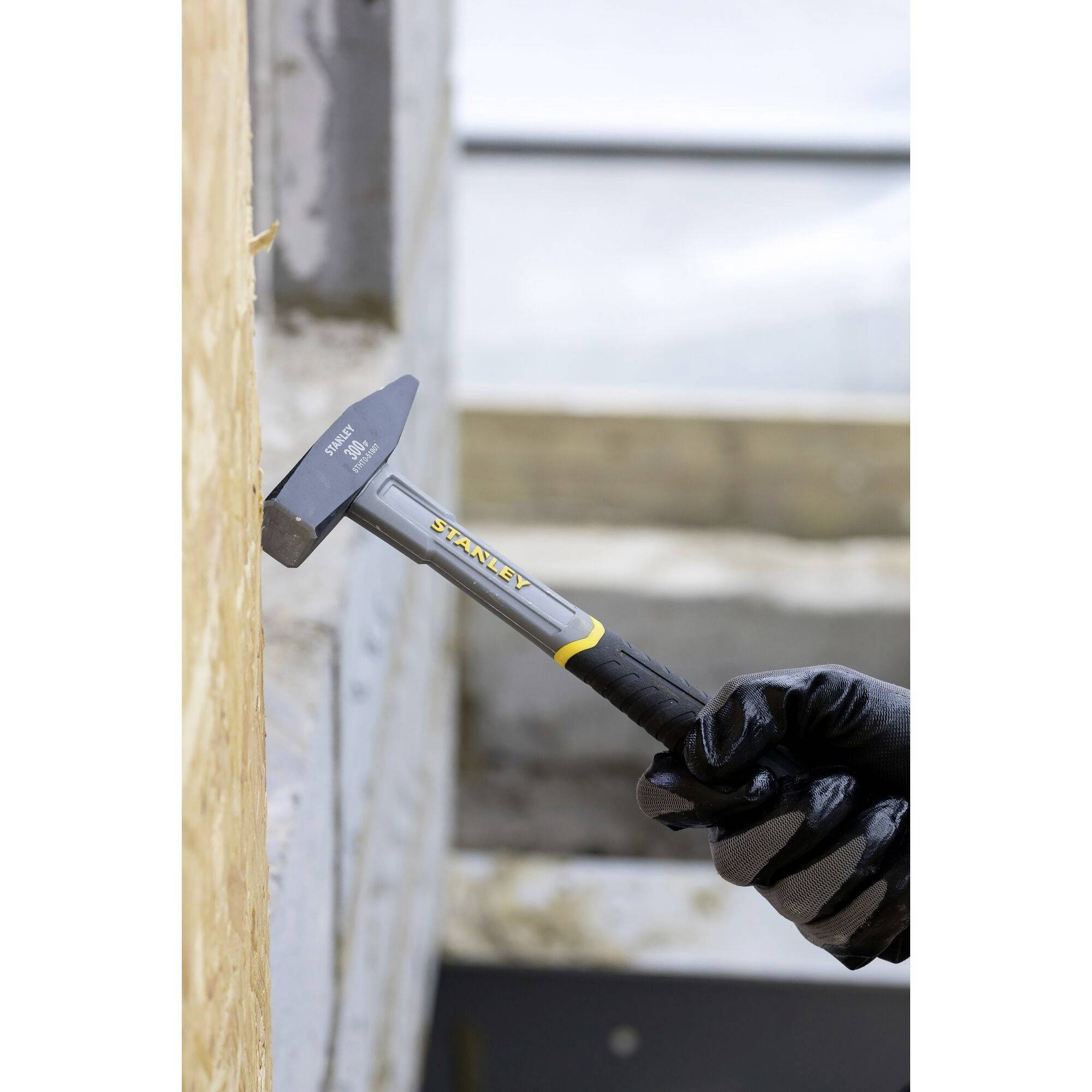 Eine Person mit schwarzen Handschuhen verwendet einen Zimmermannshammer, um ein Holzbrett zu bearbeiten.