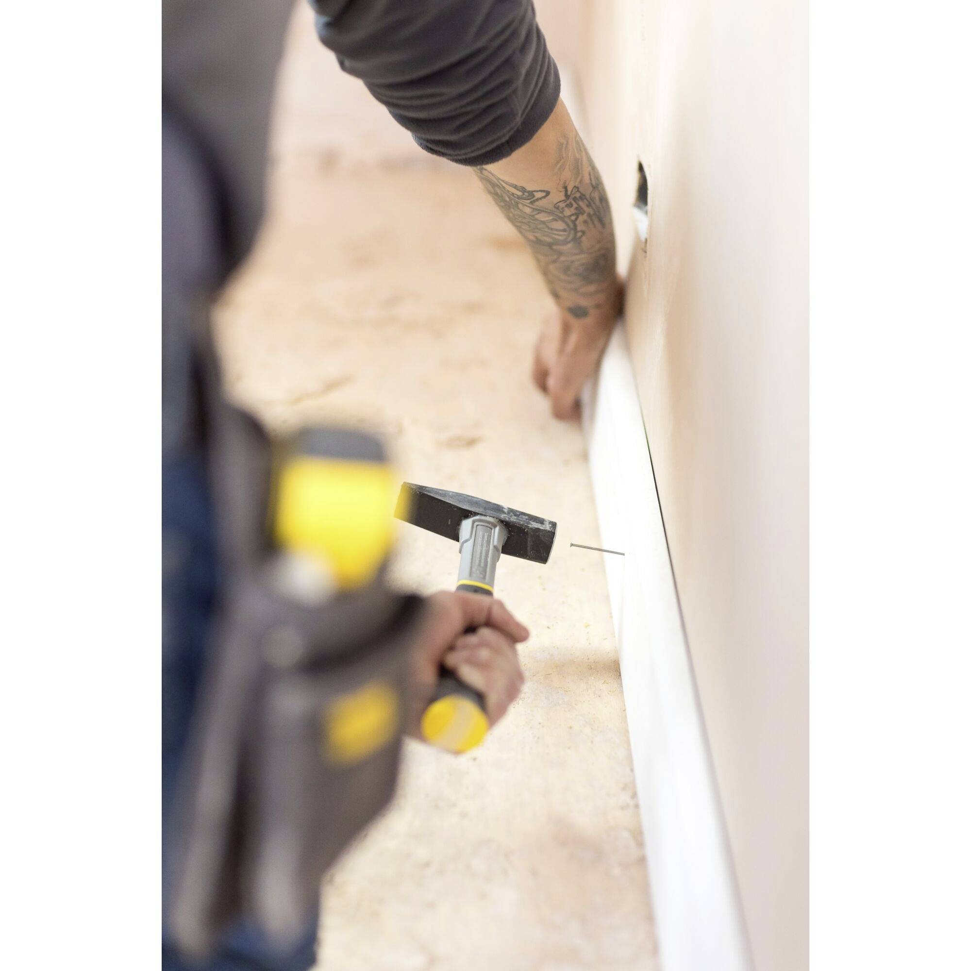 Ein Handwerker befestigt weiße Fußleisten an einer Wand. Links sind Werkzeuge am Gürtel sichtbar.