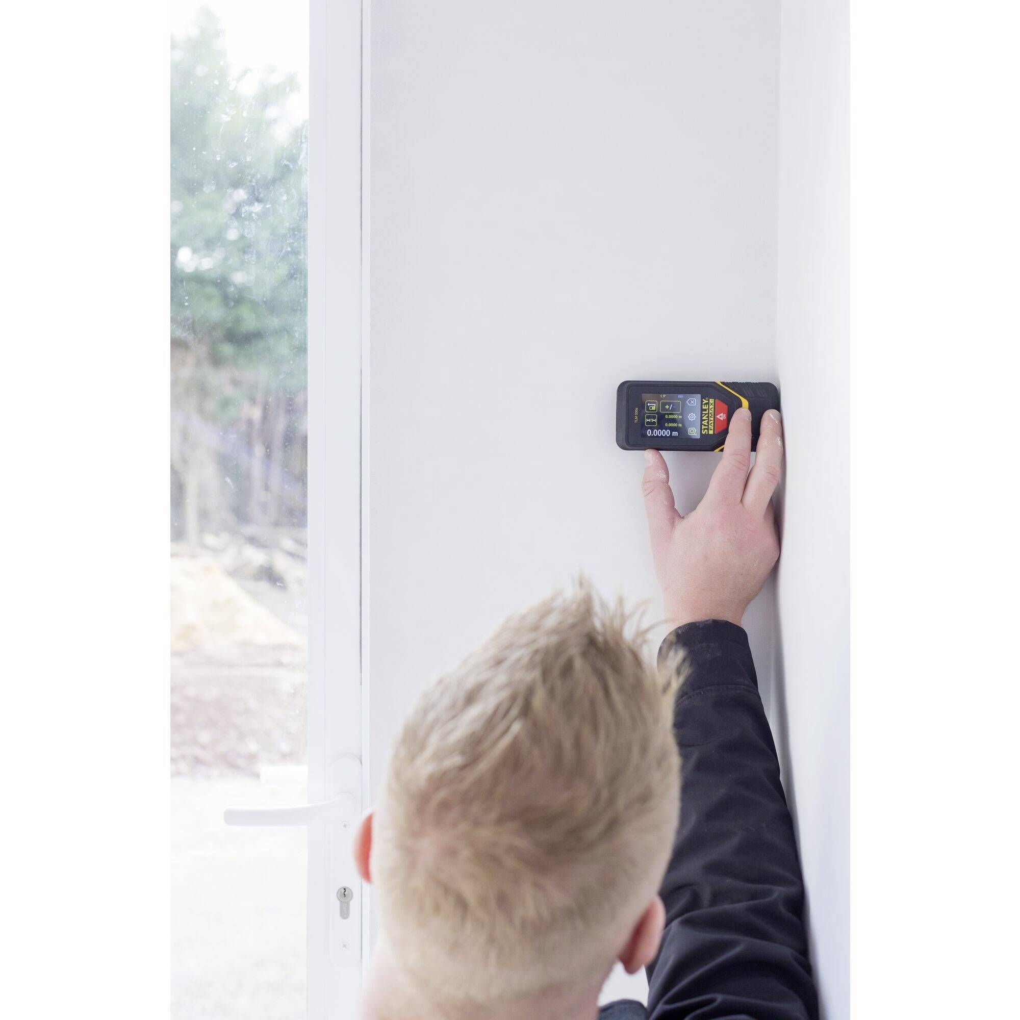 Eine Person misst mit einem digitalen Gerät die Entfernung an einer weißen Wand, umgeben von einem Fensterrahmen und Blick nach draußen.