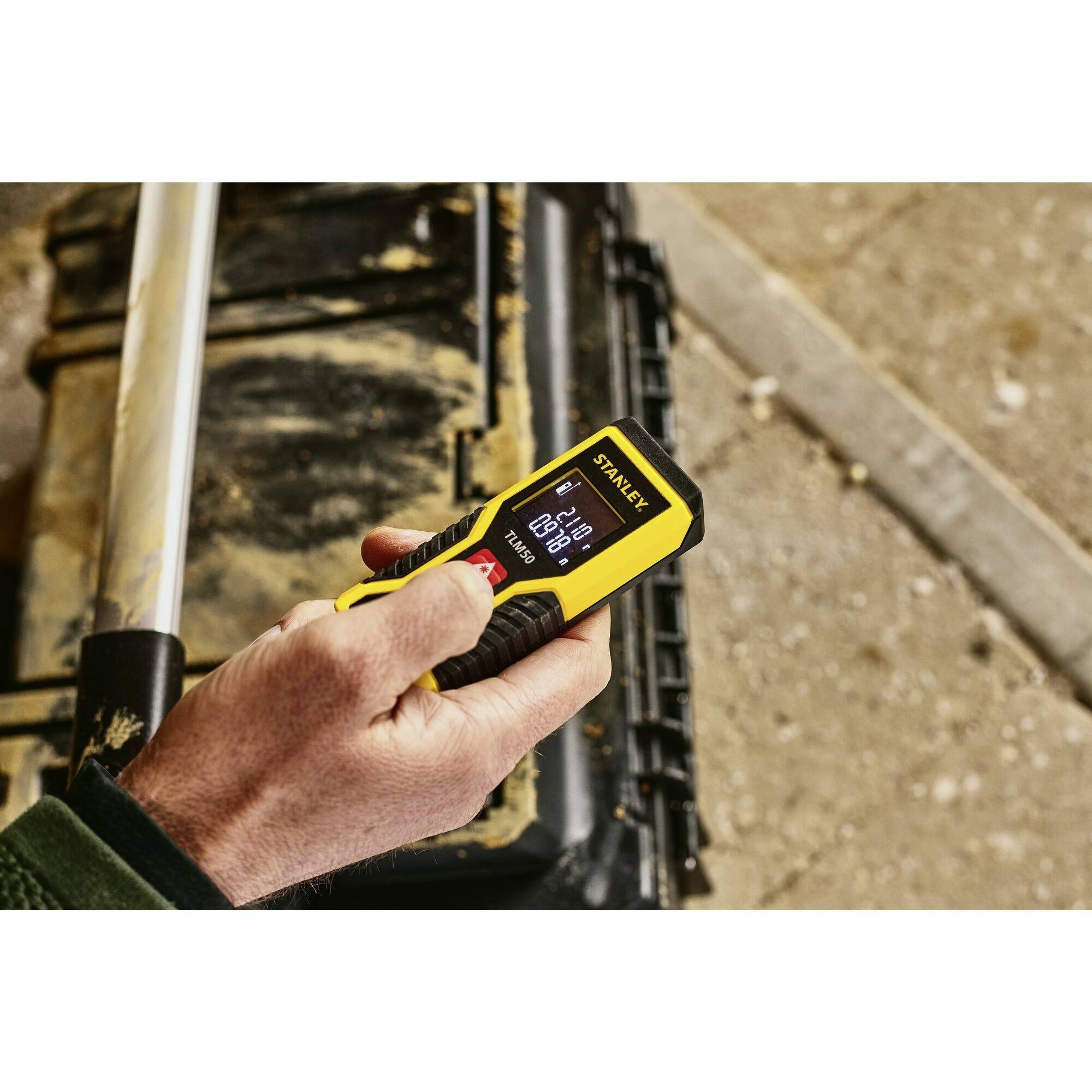 Eine Person benutzt ein gelbes digitales Laser-Entfernungsmessgerät, um eine Distanz von 4,234 Metern zu messen. Im Hintergrund ist eine Werkzeugkiste.