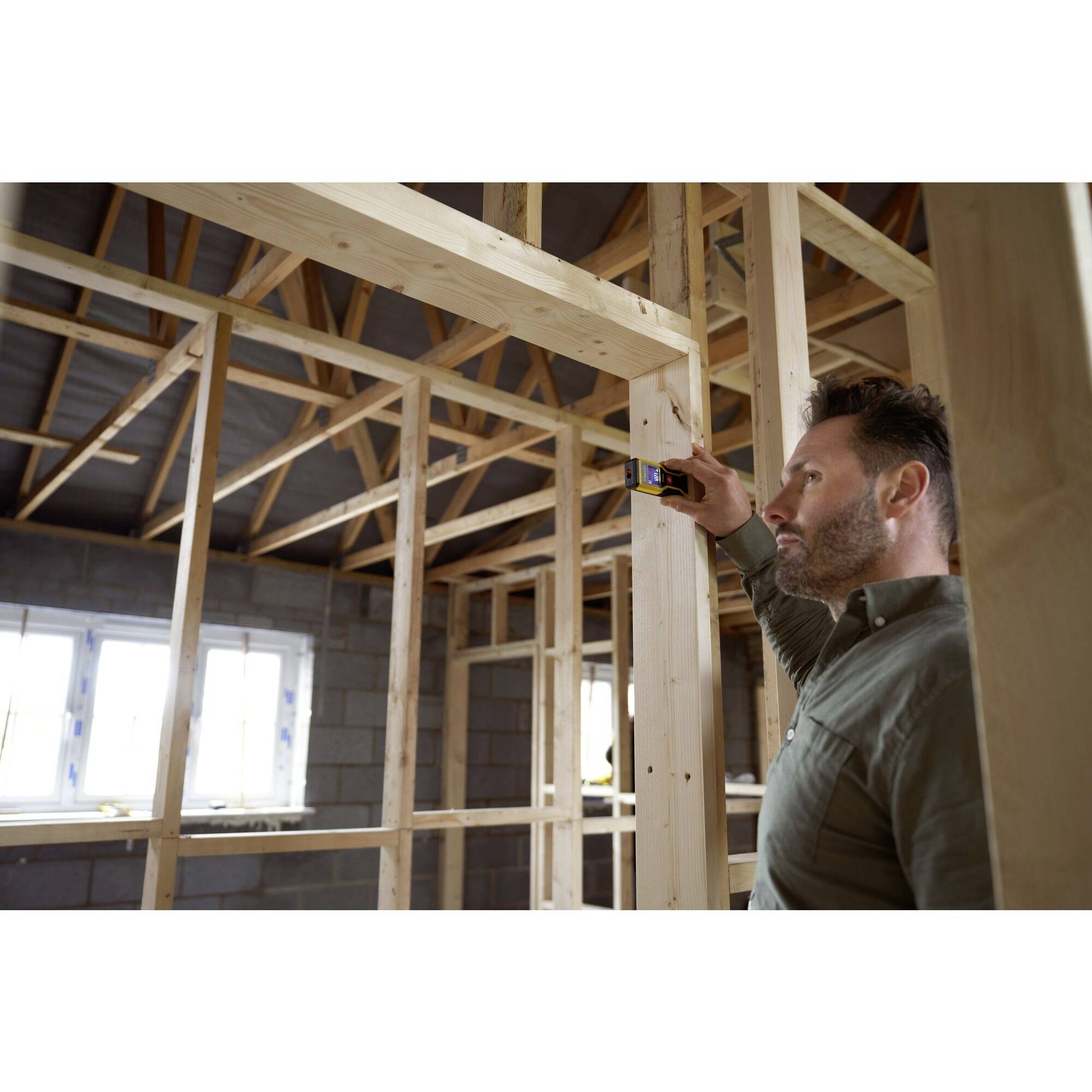 Ein Mann misst mit einem Lasermessgerät den Abstand in einem im Bau befindlichen Holzrahmengebäude.