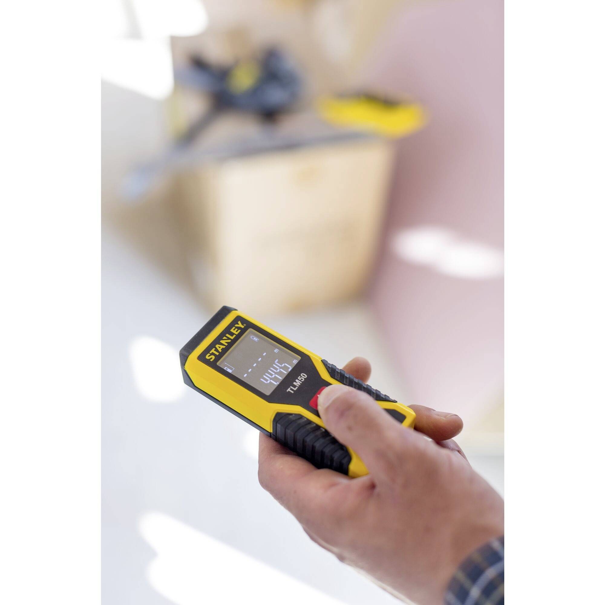 Eine Hand hält einen gelben digitalen Entfernungsmesser der Marke Staley, der eine Messdistanz von 444 mm anzeigt. Im Hintergrund verschwommenes Werkzeug.