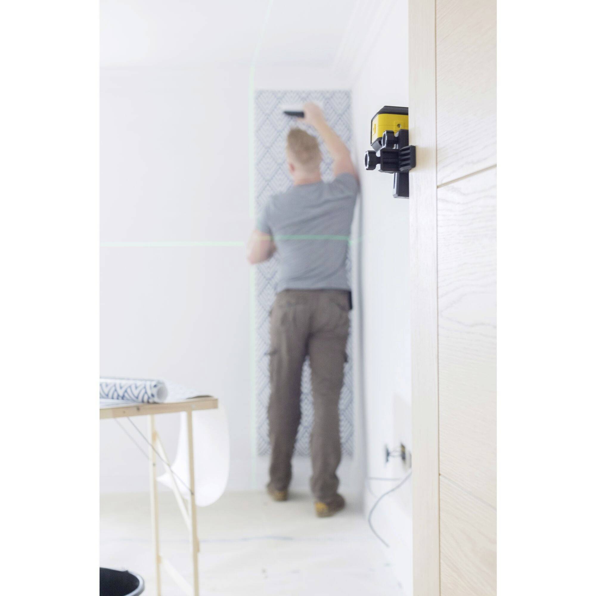 Eine Person tapeziert eine Wand, während ein Lasergerät an der gegenüberliegenden Wand Linien für Präzision projiziert.