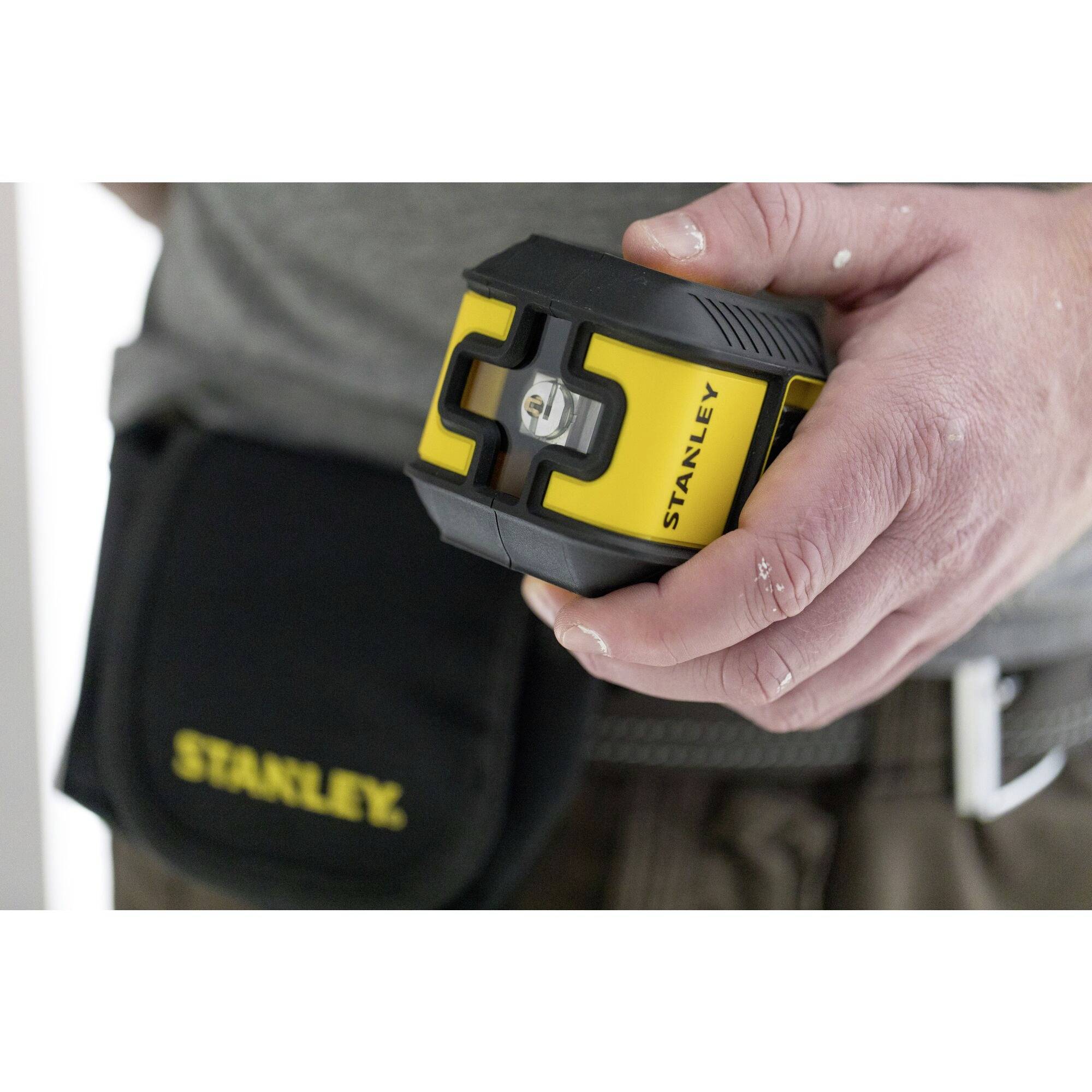 Eine Person hält ein gelbes und schwarzes Laser-Nivelliergerät in der Hand, mit einer Gürteltasche im Hintergrund.