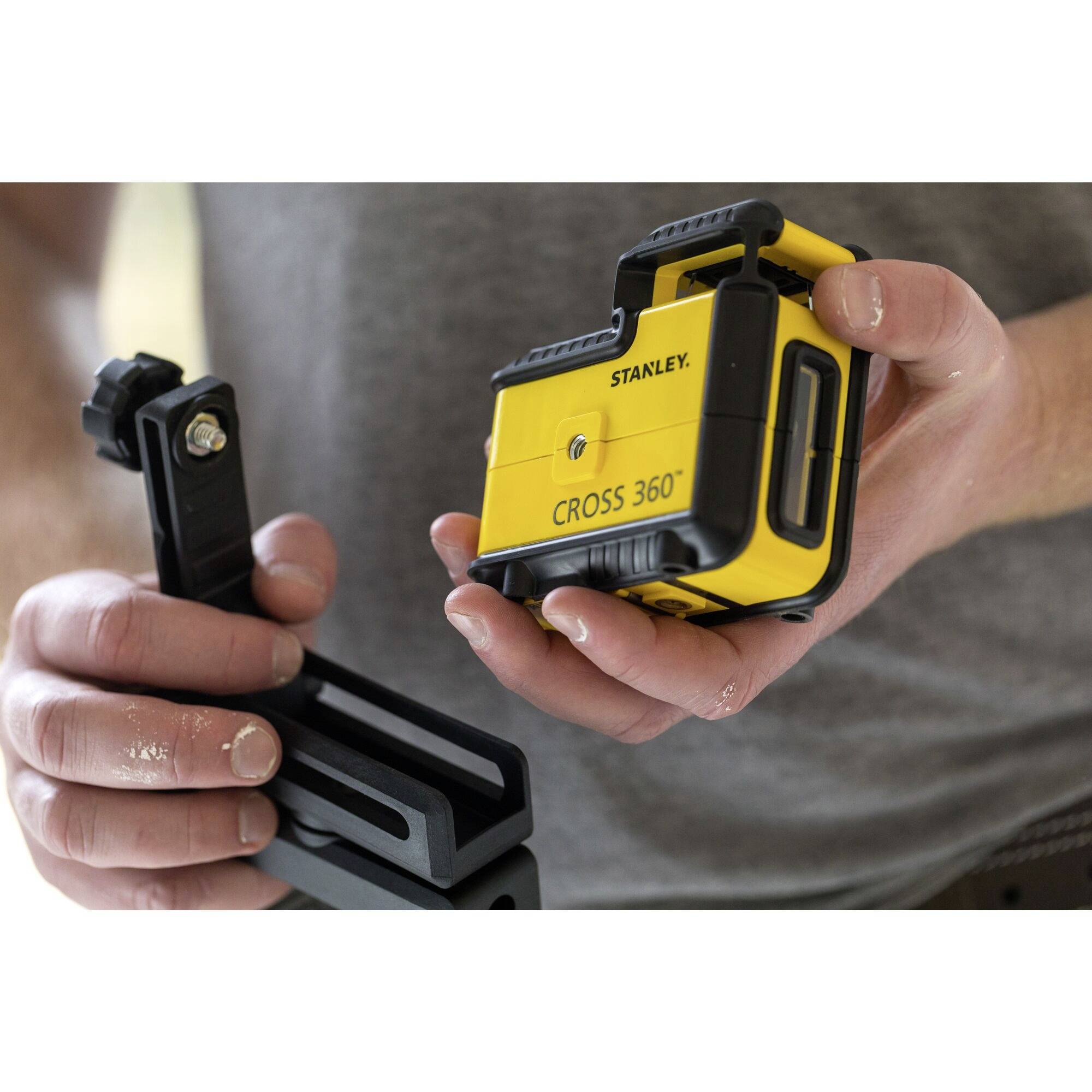 Eine Person hält ein gelbes Stanley Cross 360° Laser-Nivelliergerät in der rechten Hand und eine Halterung in der linken.