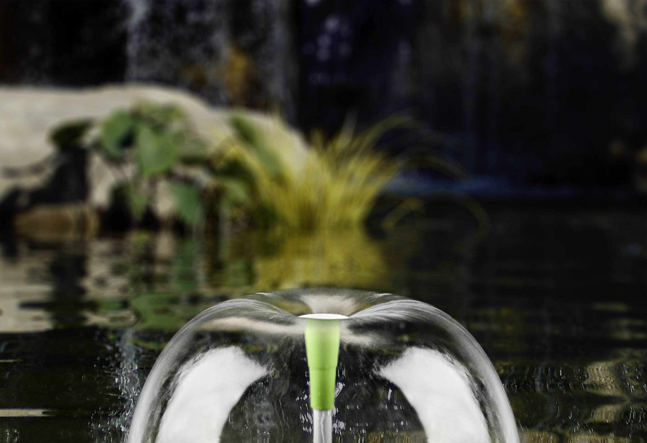 Ein Springbrunnen im Wasser bildet eine klare, halbkugelförmige Kuppel. Im Hintergrund sind Pflanzen und ein Wasserfall sichtbar.