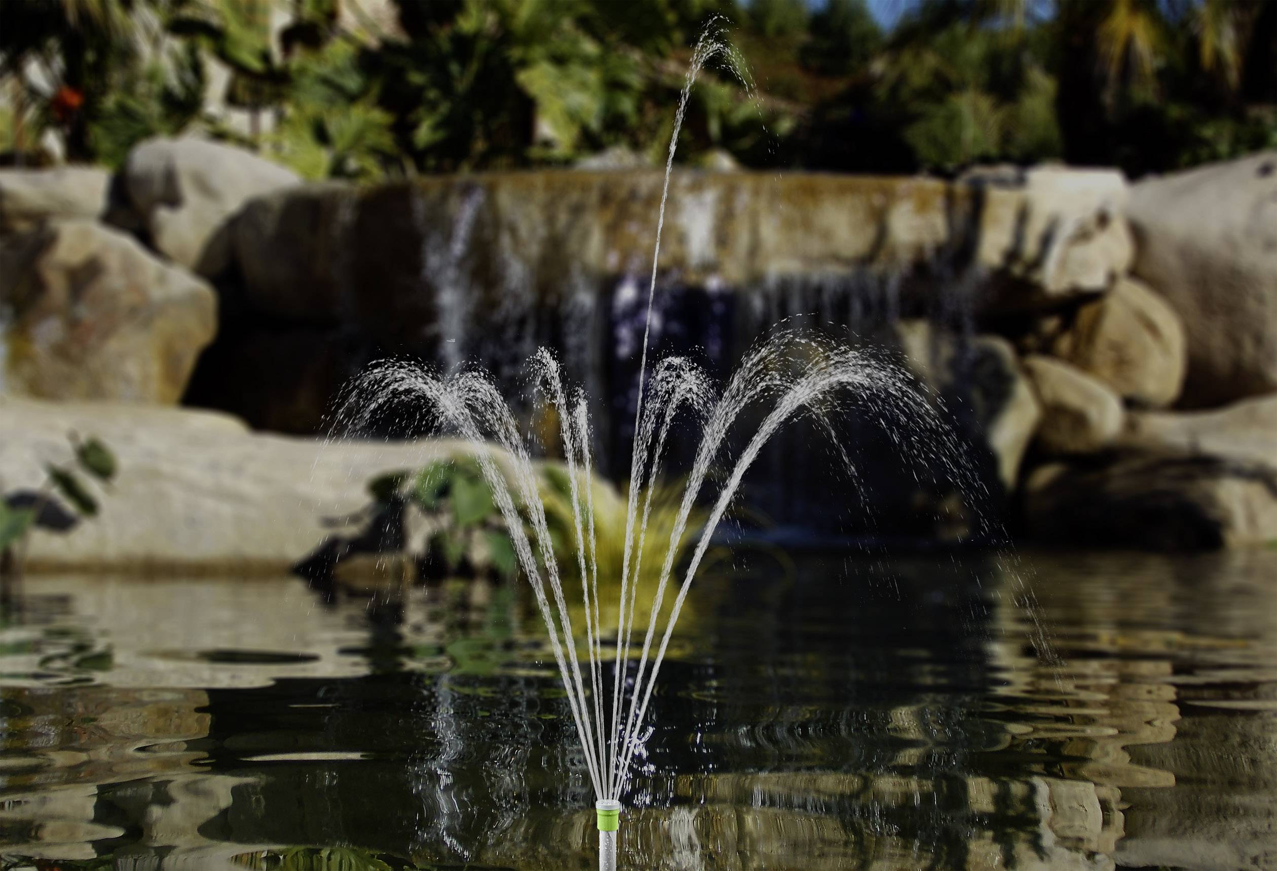 Springbrunnen in einem Teich mit einem Wasserfall im Hintergrund, umgeben von Felsen und Palmen.