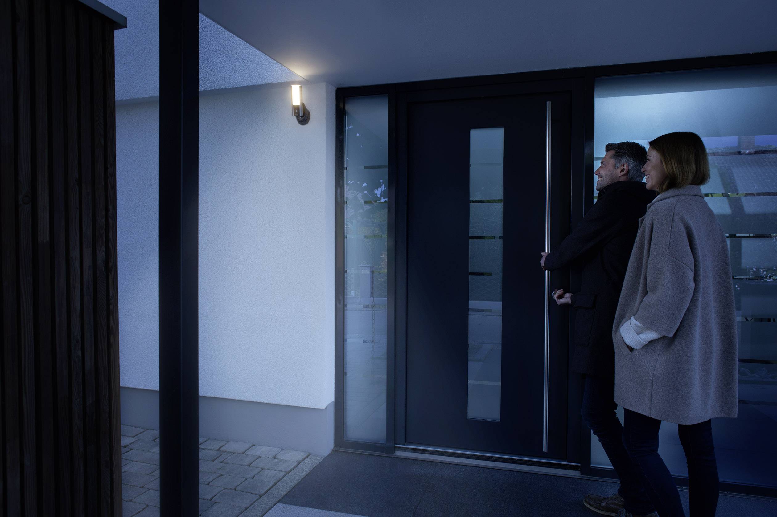 Ein Mann und eine Frau betreten bei Dämmerung ein modernes Haus durch die Eingangstür. Außenbeleuchtung erhellt den Eingangsbereich.