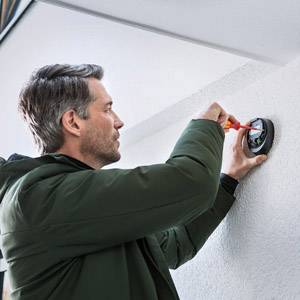 Ein Mann in grüner Jacke installiert ein rundes, elektronisches Gerät an einer weißen Wand. Er hält einen Schraubenzieher in der Hand.