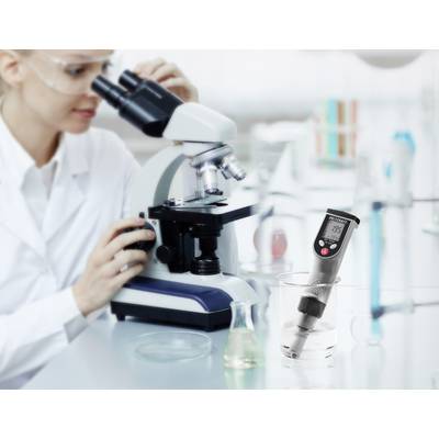 Eine Wissenschaftlerin betrachtet durch ein Mikroskop eine Probe. Ein digitales pH-Meter misst den pH-Wert einer Flüssigkeit daneben.