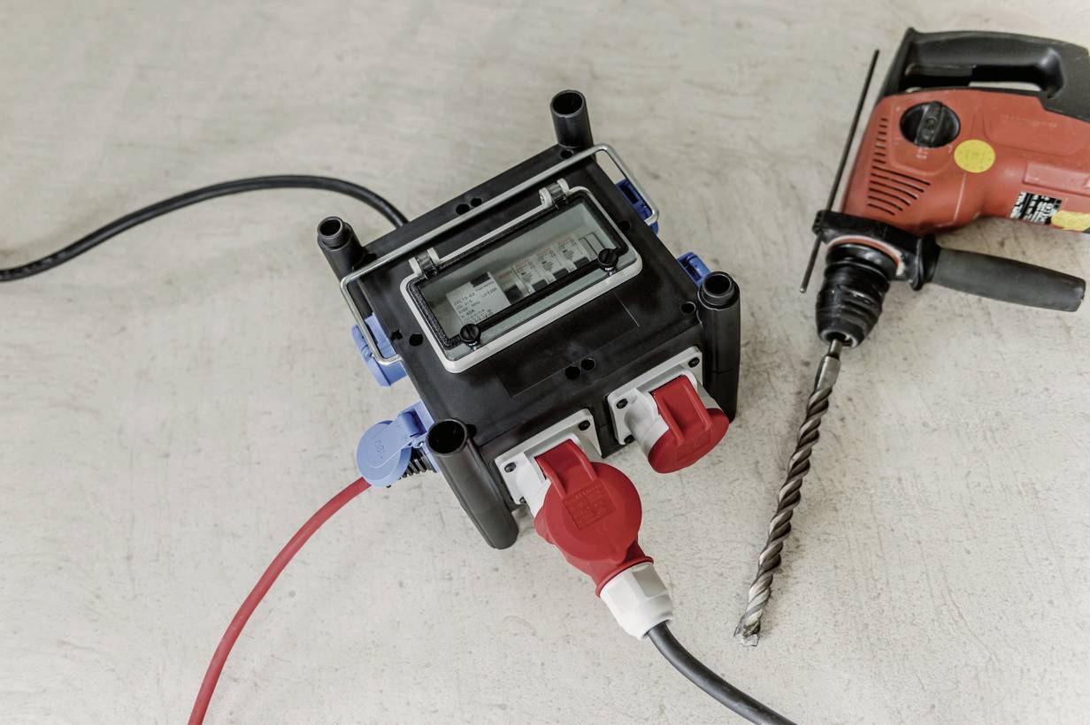 Eine Stromverteilerbox mit mehreren Steckdosen liegt auf einem Betonboden neben einem elektrischen Bohrhammer mit langem Bohrer.