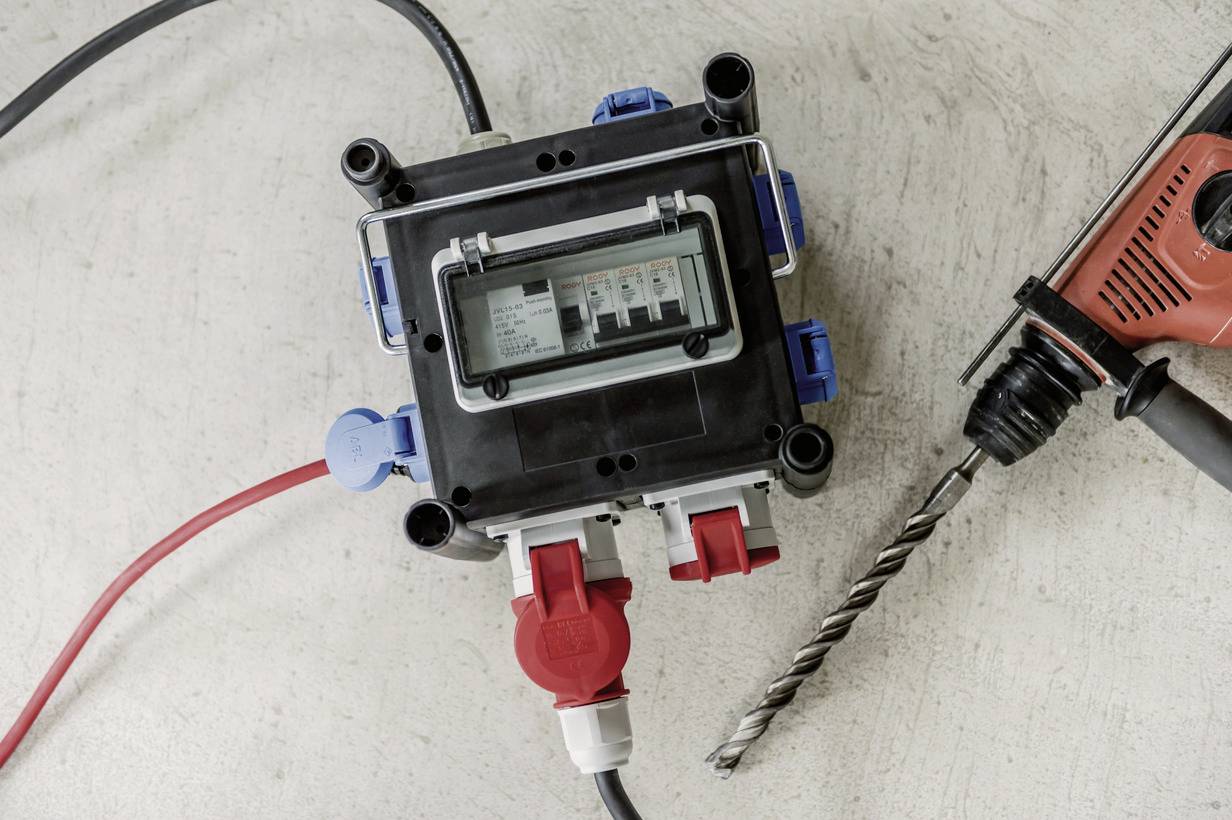 Eine schwarze Verteilerbox mit mehreren Anschlüssen liegt auf einem Betonboden. Daneben ein roter Bohrhammer mit Bohrer.