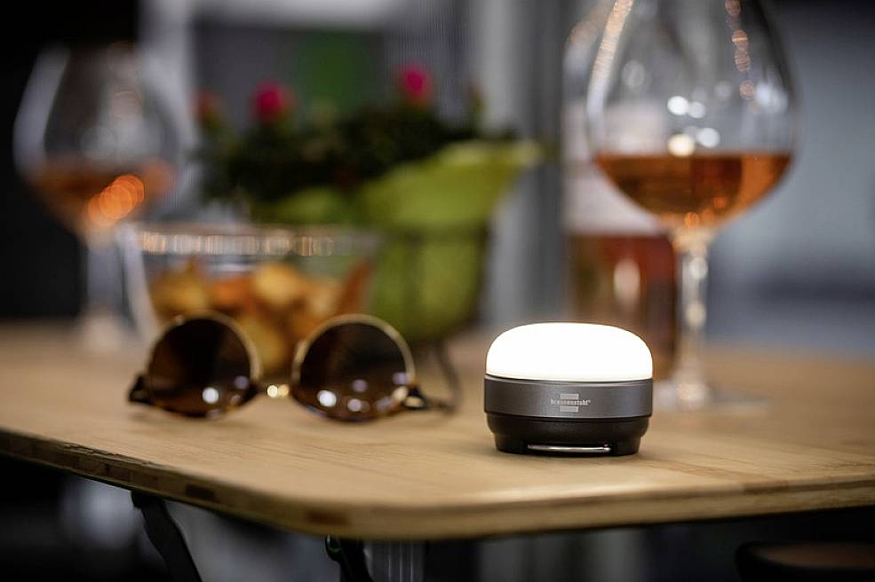 Ein kleiner, leuchtender LED-Lampe steht auf einem Holztisch. Im Hintergrund sind Sonnenbrille, Getränke und Topfpflanze zu sehen.