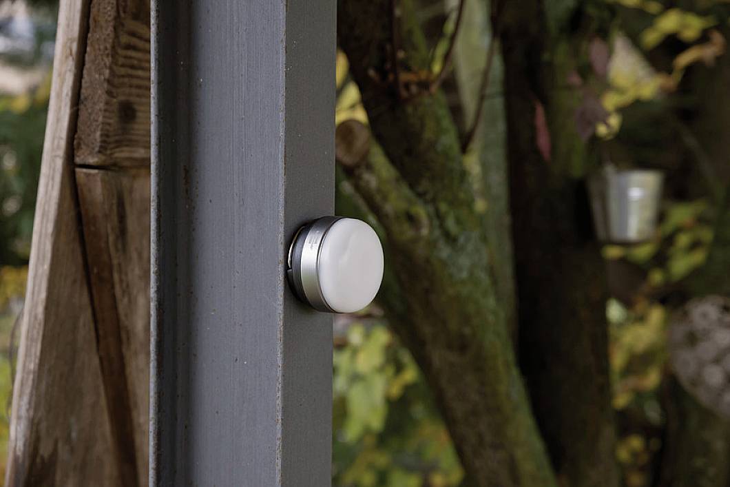 Ein weißer Rundsensor an einem Türrahmen in einer natürlichen Umgebung, möglicherweise im Freien. Hintergrund zeigt Bäume und Laub.