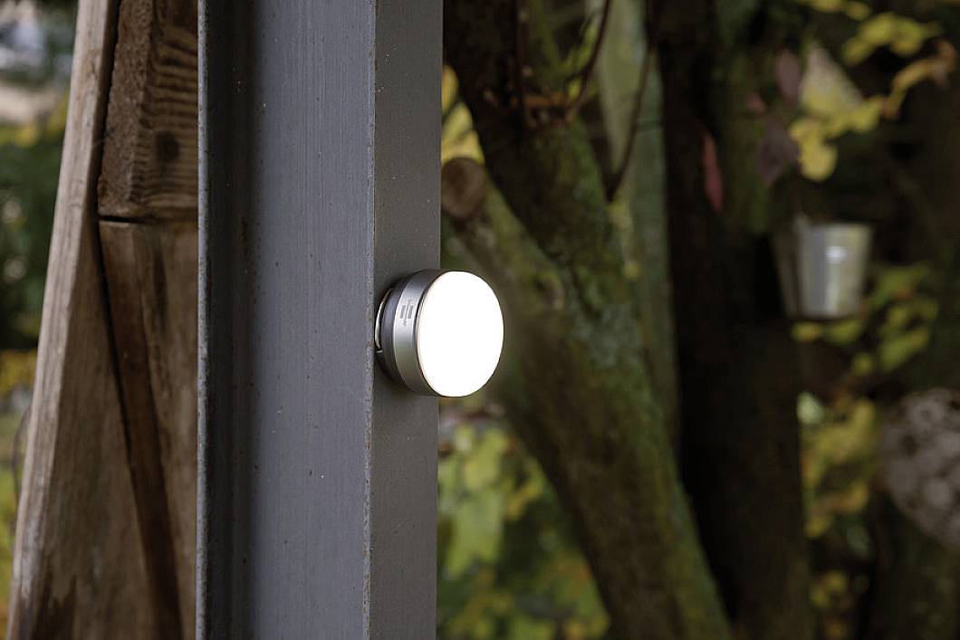 Eine runde Lampe leuchtet an einem dunklen Holzpfosten im Freien. Im Hintergrund sind unscharfe Bäume zu sehen, die auf einen Wald hinweisen.