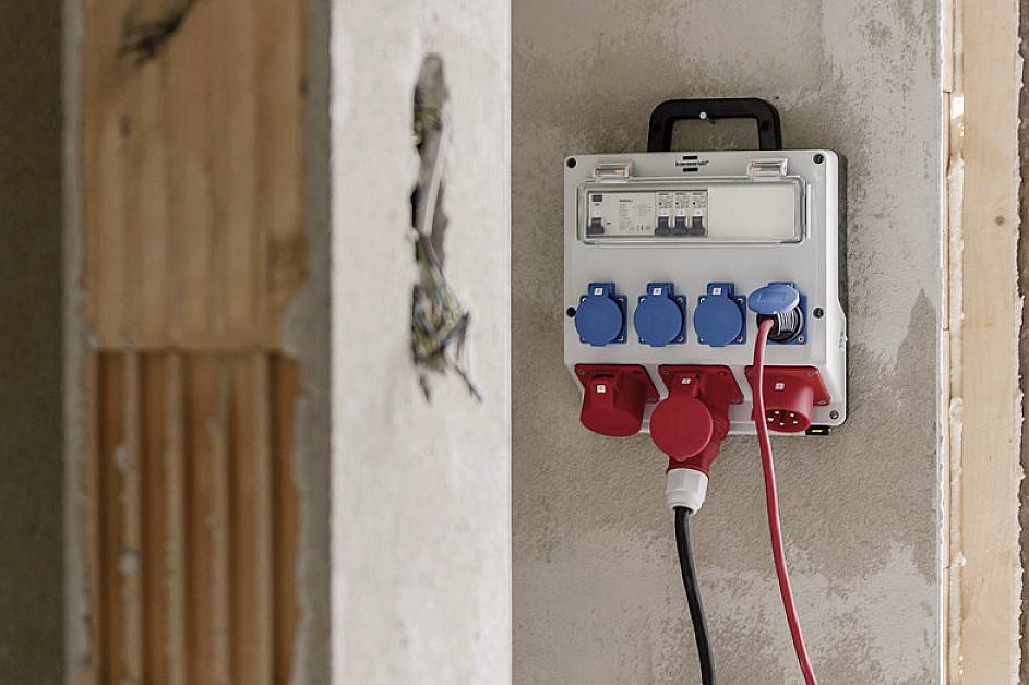 Elektrische Schalttafel an einer Wand montiert, mit mehreren Steckdosen und Kabeln. Baustellenumgebung im Hintergrund.