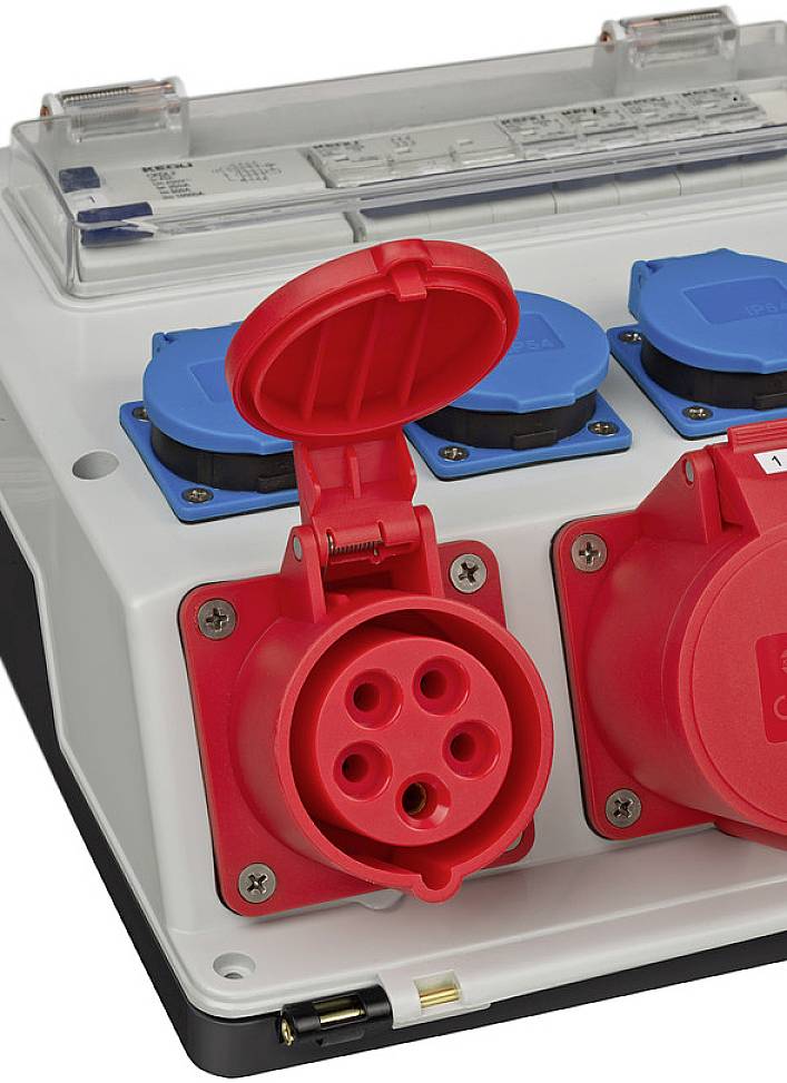 Ein graues elektrisches Anschlussfeld mit roten und blauen Steckdosen. Eine rote Abdeckung ist geöffnet, um die Anschlüsse zu zeigen.