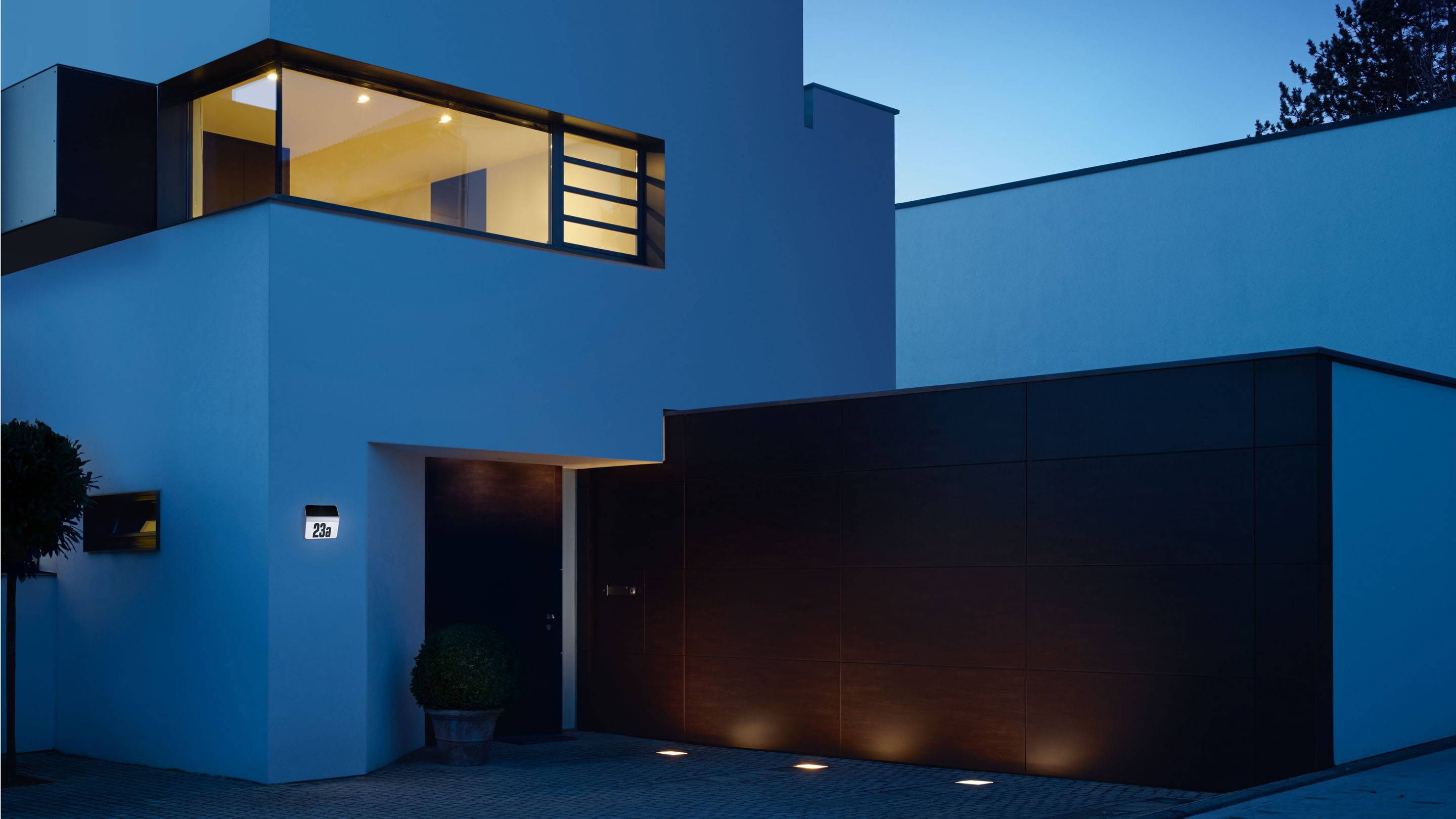 Modernes Haus bei Abenddämmerung mit beleuchtetem Innenraum und Außenleuchten; klare, geometrische Linien und dunkle Holzverkleidung.