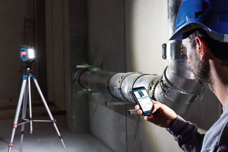 Ein Bauarbeiter mit Schutzhelm betrachtet ein Smartphone, während er vor einem Rohr steht. Eine Leuchte auf einem Stativ ist ihm zugewandt.
