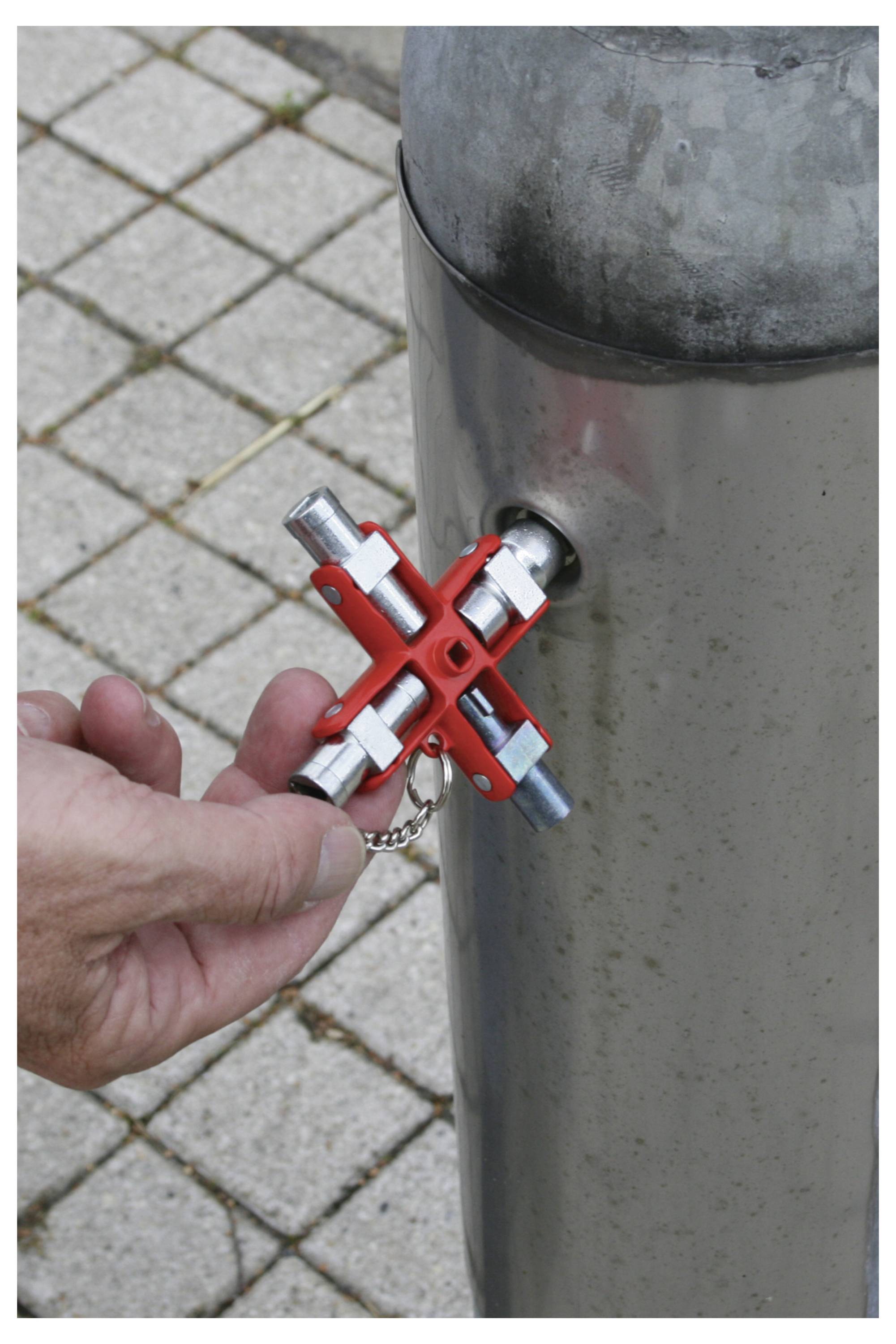 Eine Person benutzt einen roten Vierkantschlüssel, um einen Wasserhahn zu öffnen, der an einem Metallpoller auf einem gepflasterten Gehweg angebracht ist.