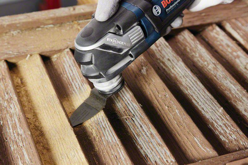 Eine Hand hält ein elektrisches Multifunktionswerkzeug mit einem Sägeblattaufsatz, das altes Holz zwischen Latten abschleift und bearbeitet.