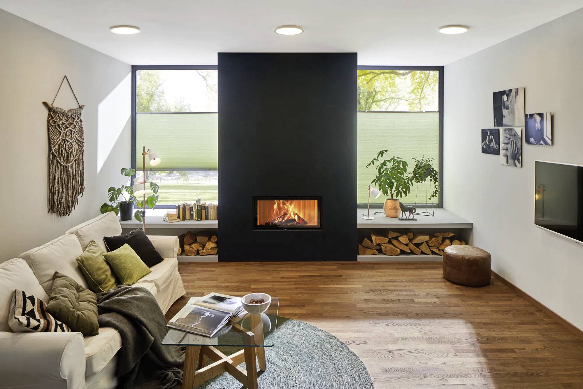 Ein modernes Wohnzimmer mit einem weißen Sofa, bunten Kissen, einem zentralen schwarzen Kamin, Holzboden und großen Fenstern mit Pflanzen.