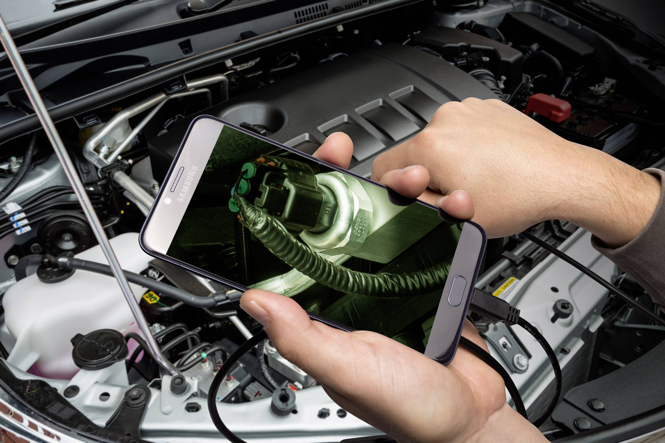Eine Person hält ein Smartphone über einen geöffneten Motorraum und zeigt ein Bild eines Teils des Motors auf dem Bildschirm.