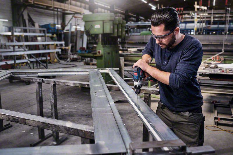 Ein Mann in Schutzbrille benutzt eine Bohrmaschine, um Metallrahmen in einer Werkstatt zu befestigen. Industrielle Maschinen im Hintergrund.