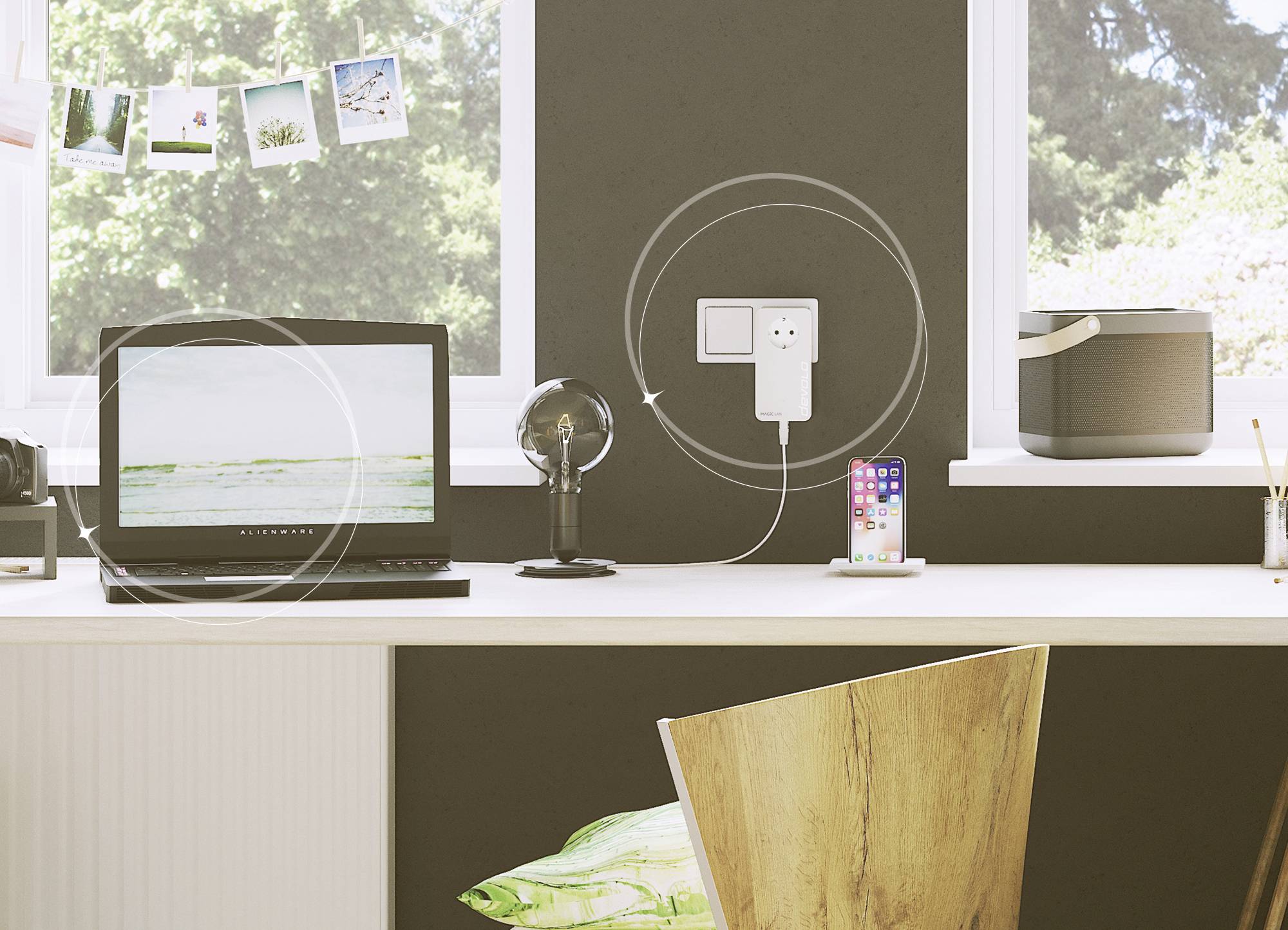 Ein Schreibtisch mit einem Laptop, einer Lampe, einem Mehrfachstecker an der Wand, einem Smartphone und einem Lautsprecher. Fenster im Hintergrund zeigen draußen Bäume.