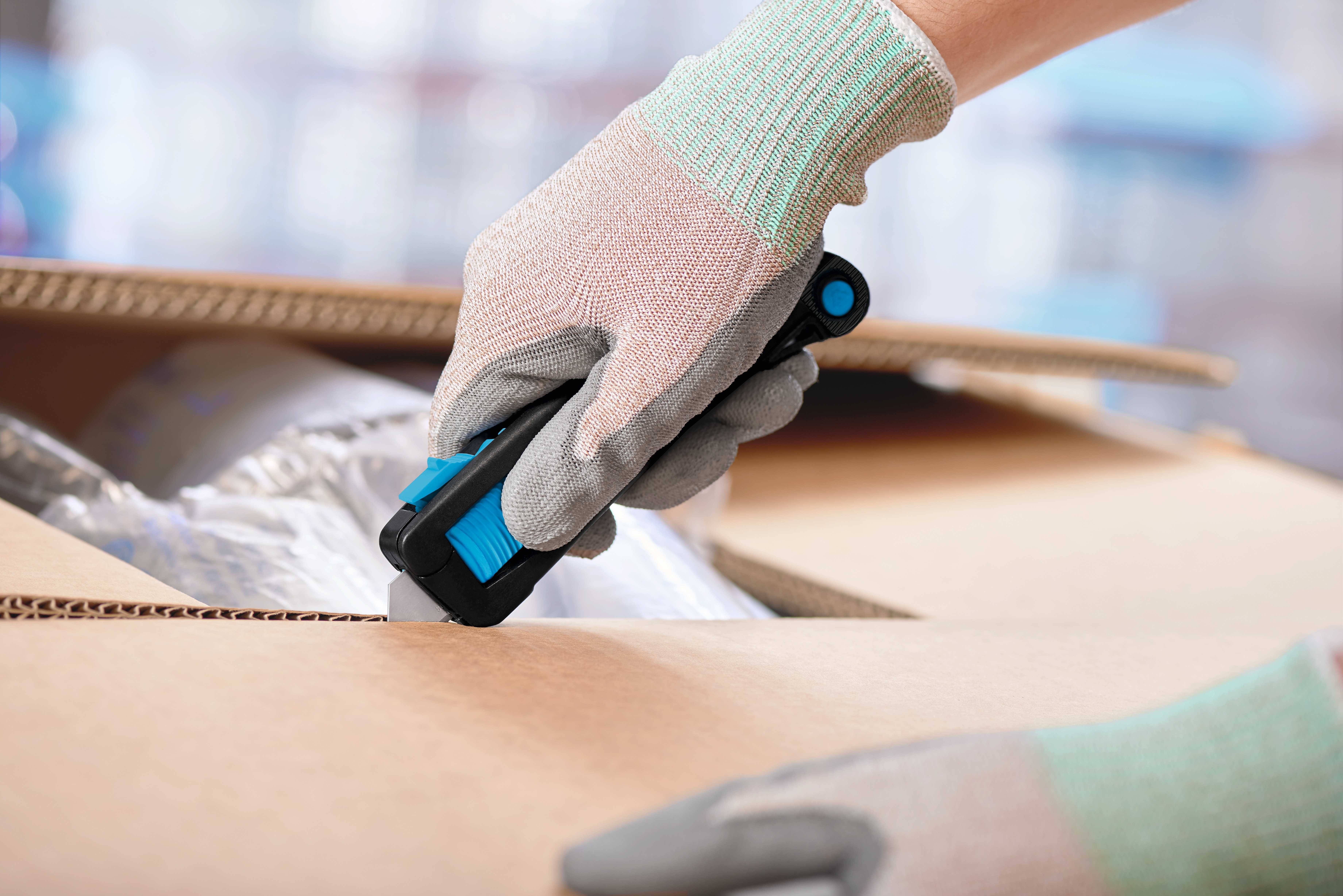 Eine behandschuhte Hand öffnet mit einem blauen Cutter-Messer einen Karton. Die Szene zeigt eine vorsichtige Arbeitsweise beim Auspacken.