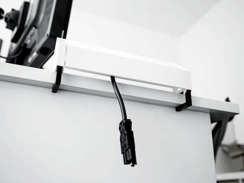 Eine weiße Kabelbox, die unter einem Schreibtisch befestigt ist, mit einem schwarzen Netzkabel, das herunterhängt und eine organisierte Arbeitslösung zeigt.