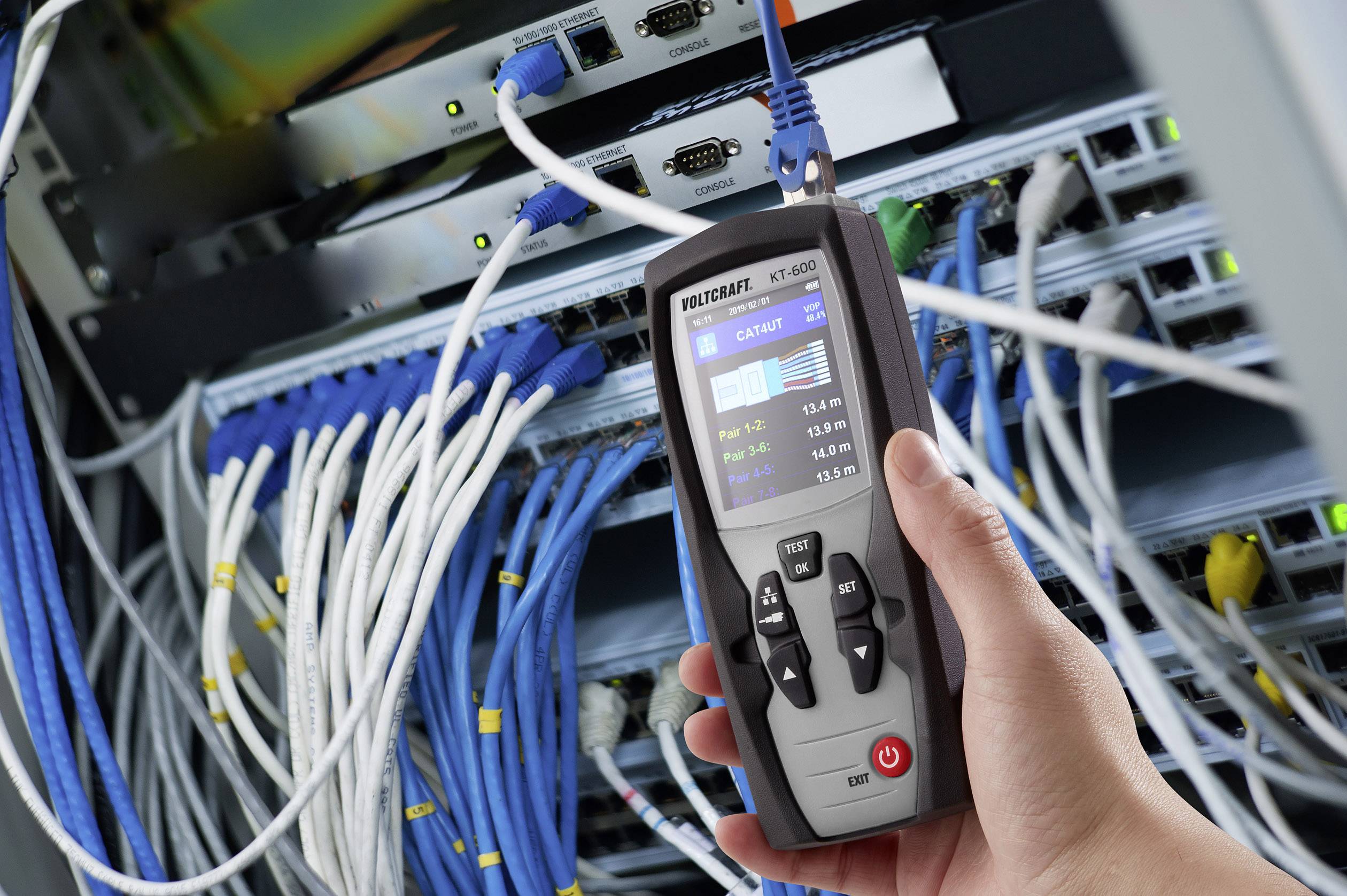 Eine Hand hält ein Netzwerkanalysegerät vor einem Serverschrank voller verkabelter Netzwerkgeräte, um Verbindungen zu überprüfen.